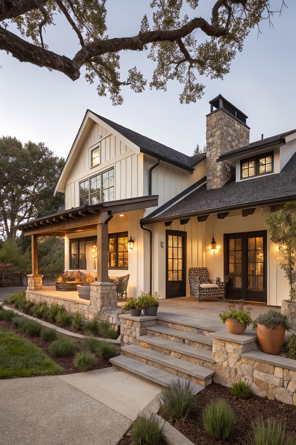 White board-and-batten ranch house exterior with gabled roof, black-trimmed windows, stone chimney and pillars supporting a covered porch, steps with stone walls, grasses and potted plants in landscape beds, at dusk.