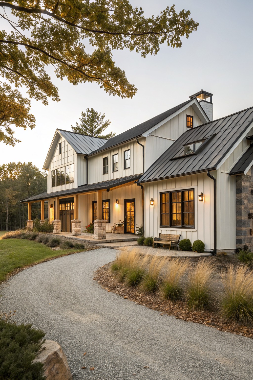 White board-and-batten sided modern farmhouse ranch house with dark metal gabled roofs, wraparound porch supported by stone columns, large windows, gravel driveway curving in front, ornamental grasses, and trees at dusk.
