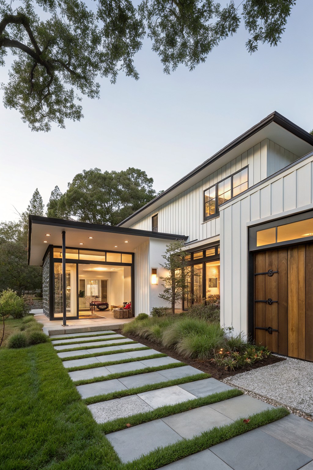 Exterior of a modern white farmhouse ranch home at dusk with board-and-batten siding, black-framed glass entry pavilion, wooden garage door, concrete stepping stone pathway inset with grass, ornamental grasses, and surrounding oak trees.