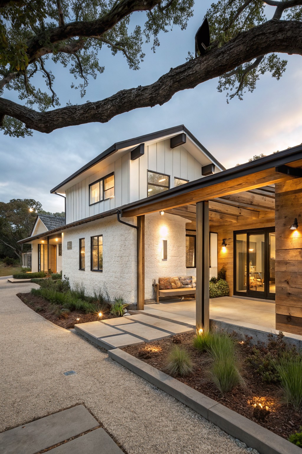 Side exterior view of a two-story modern farmhouse ranch house featuring white board-and-batten siding, black-framed windows, a covered porch with exposed wood beams and posts, built-in bench, gravel path, and low native grasses at dusk.