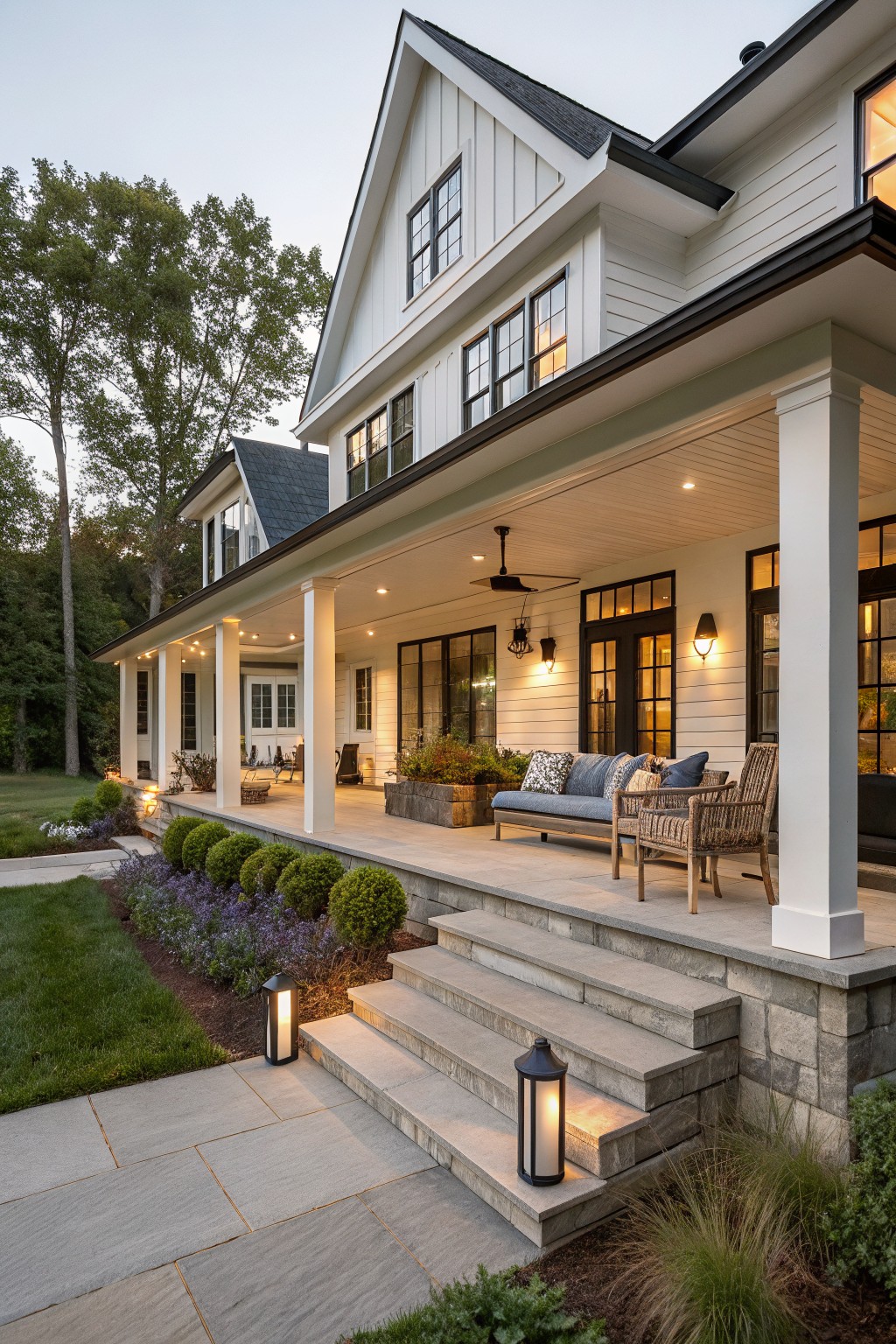 White board-and-batten sided modern farmhouse with wrap-around porch on the corner, tall white columns, black-framed windows and doors, wicker seating with blue cushions on the porch, stone steps flanked by lanterns and landscaping.