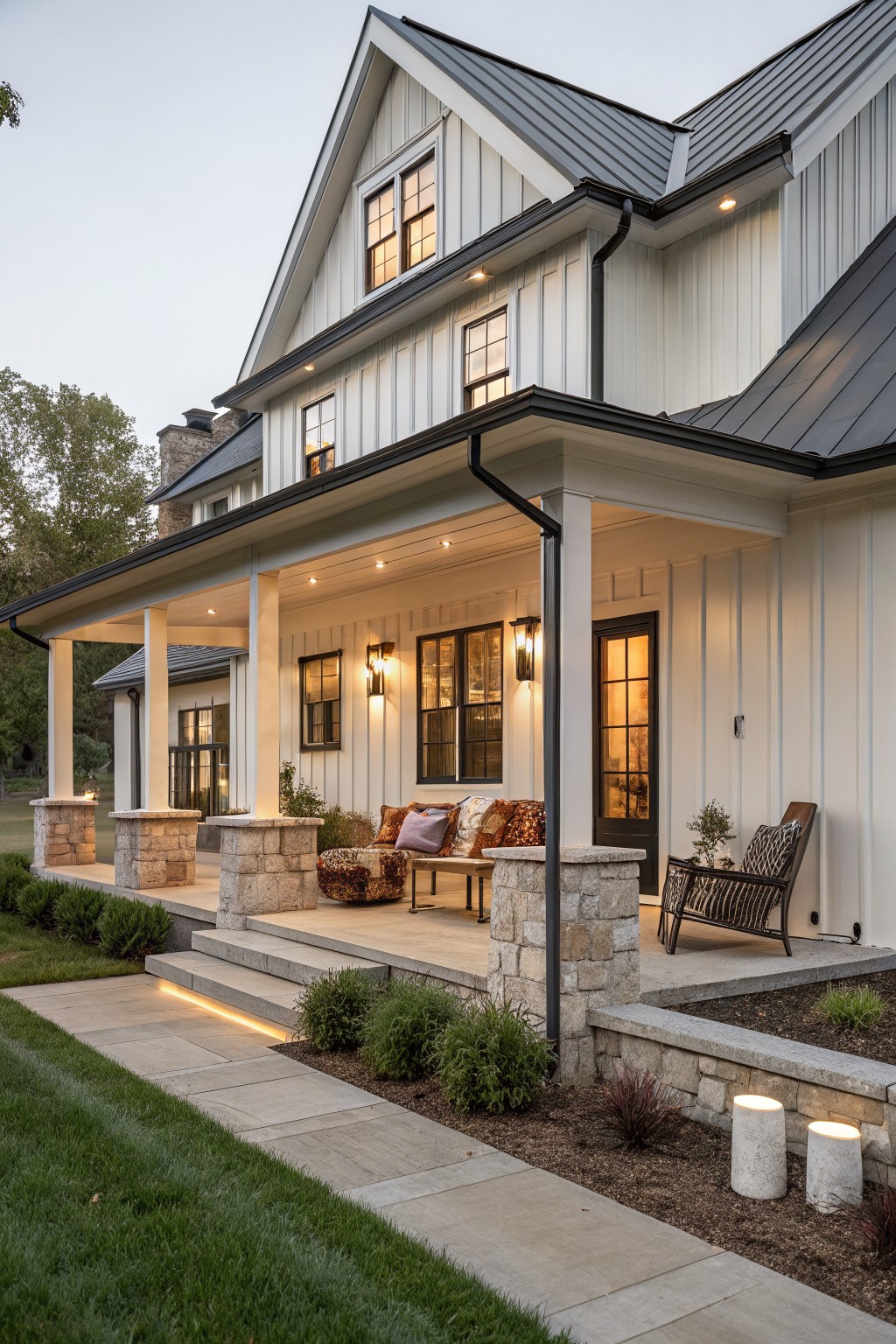 White board-and-batten sided modern farmhouse with black metal roof and trim, wraparound porch supported by stone piers, outdoor seating on porch, steps down to lit stone pathway, shrubs and grass in yard at dusk.