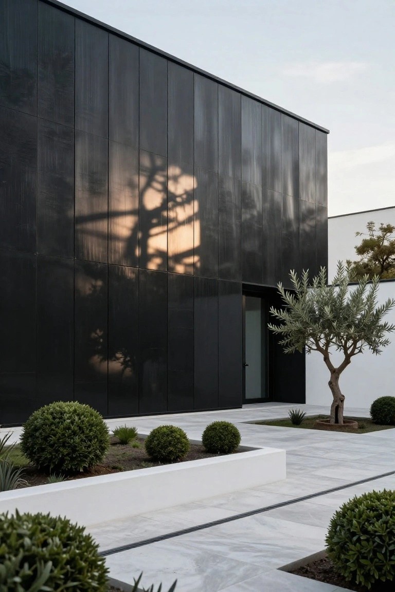 Black metal-paneled modern house facade with vertical texture, glass entry door, olive tree, boxwood shrubs, and white planters on a light stone-paved courtyard at dusk.