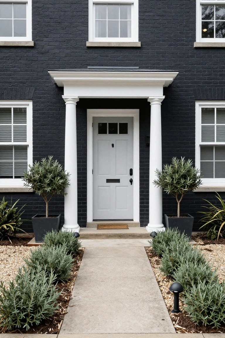 A one-story black brick house exterior featuring a white portico with columns framing a white front door, potted olive trees on either side, and a concrete path edged with lavender plants and gravel.