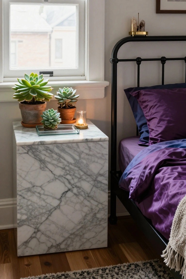 White marble cube nightstand beside black iron bed with purple satin bedding and pillows, topped with three potted succulents, glass tray, gold incense holder, lit glass candle, and crystals near a window.