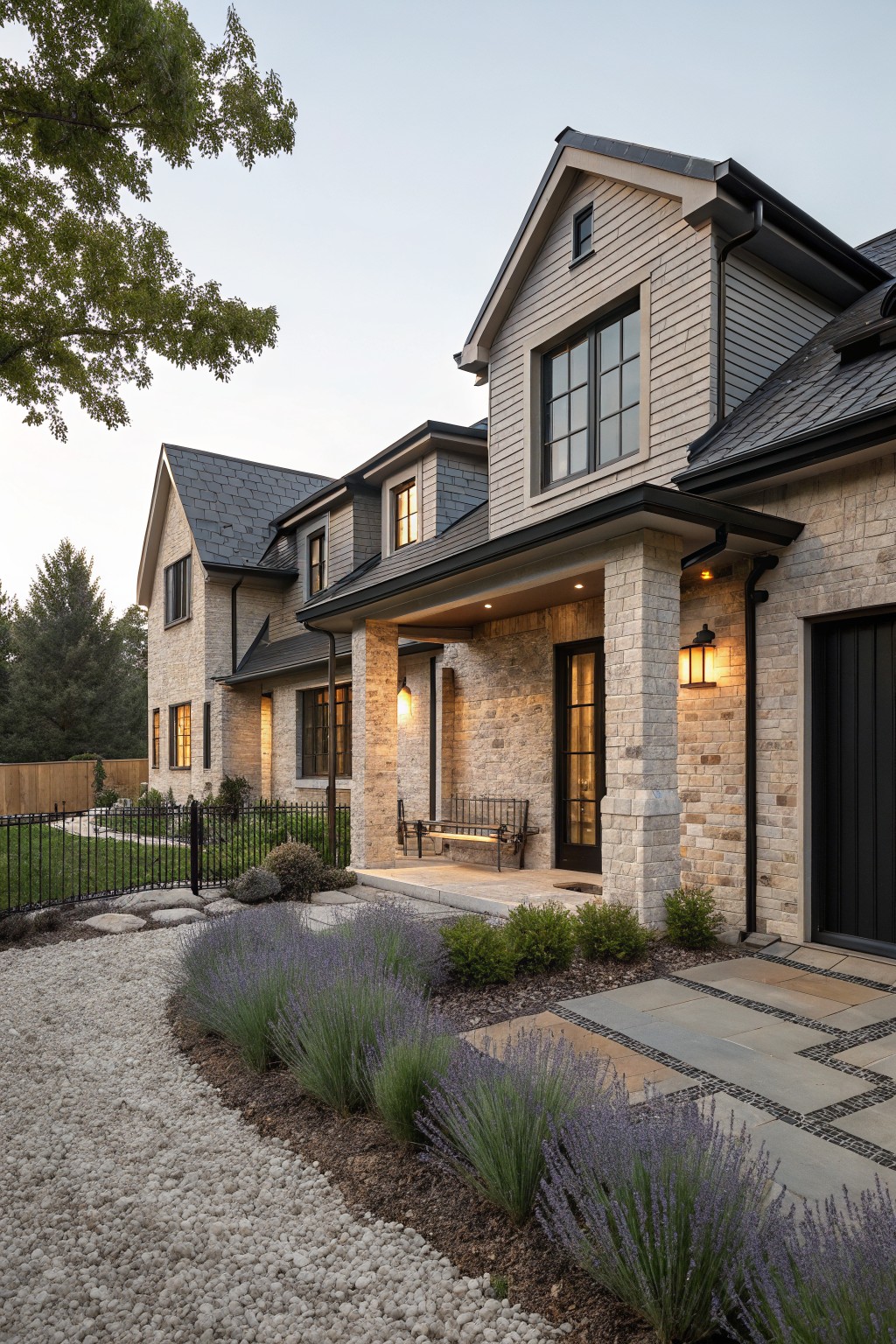 Two-story house exterior with light wood shingle siding, dark shingled roof, beige stone pillars supporting covered porch with black door and bench, black garage door, gravel path with lavender plants.