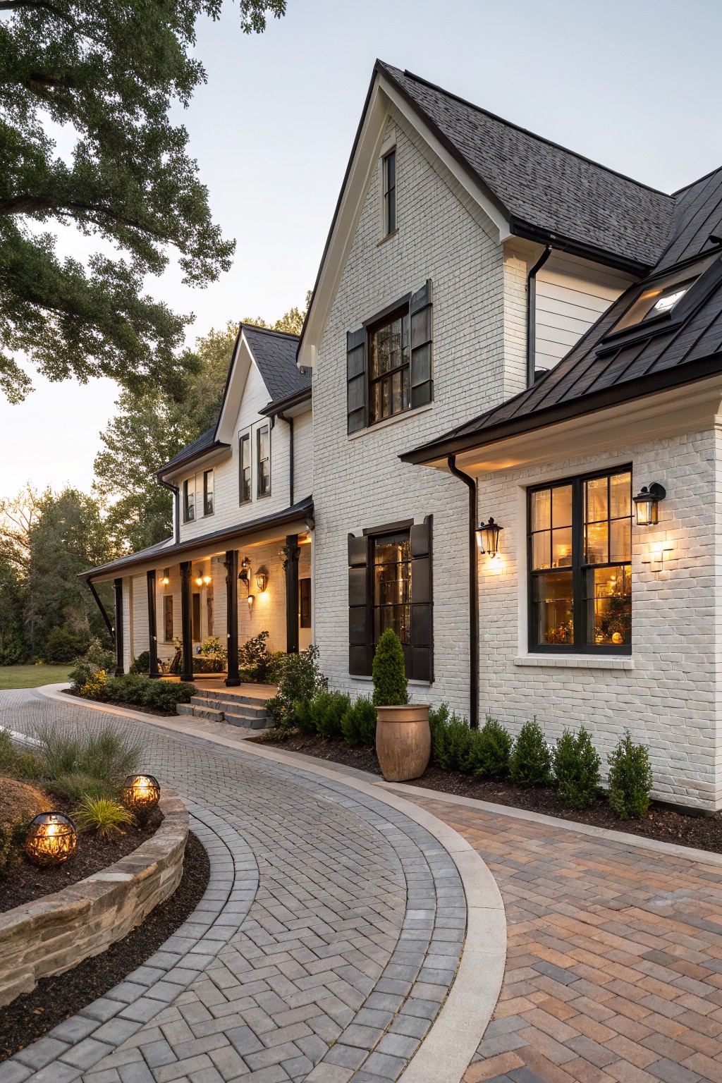 A two-story white brick farmhouse exterior featuring a black metal roof, wraparound porch with columns, black shutters, and a curved brick driveway edged with stone walls and landscaping.