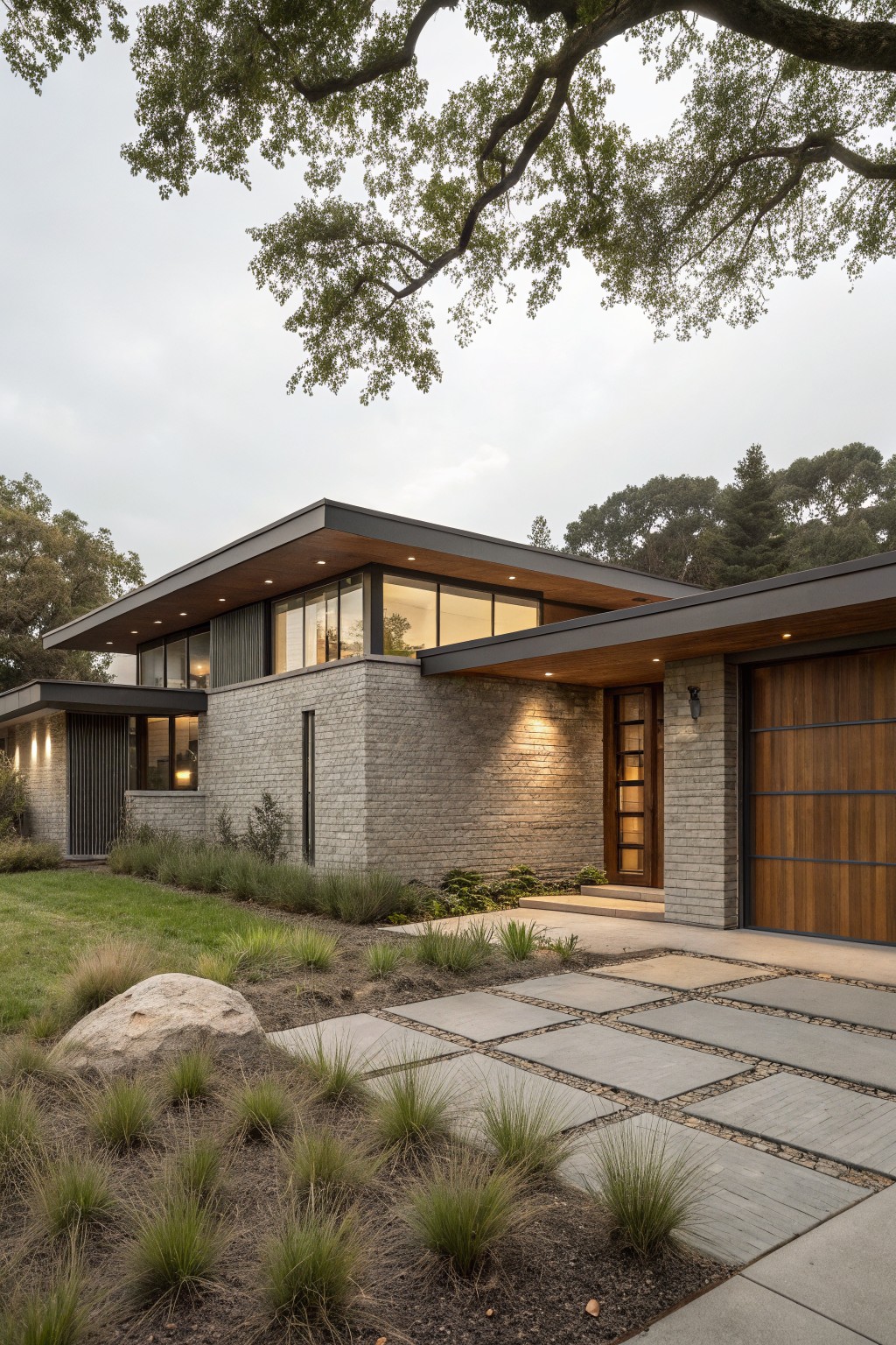Modern two-story house exterior with light gray brick walls, dark wood garage door, cantilevered dark roof overhangs, large windows, entry light, stone paver pathway, grasses, and boulder in the landscaped front yard under oak trees.