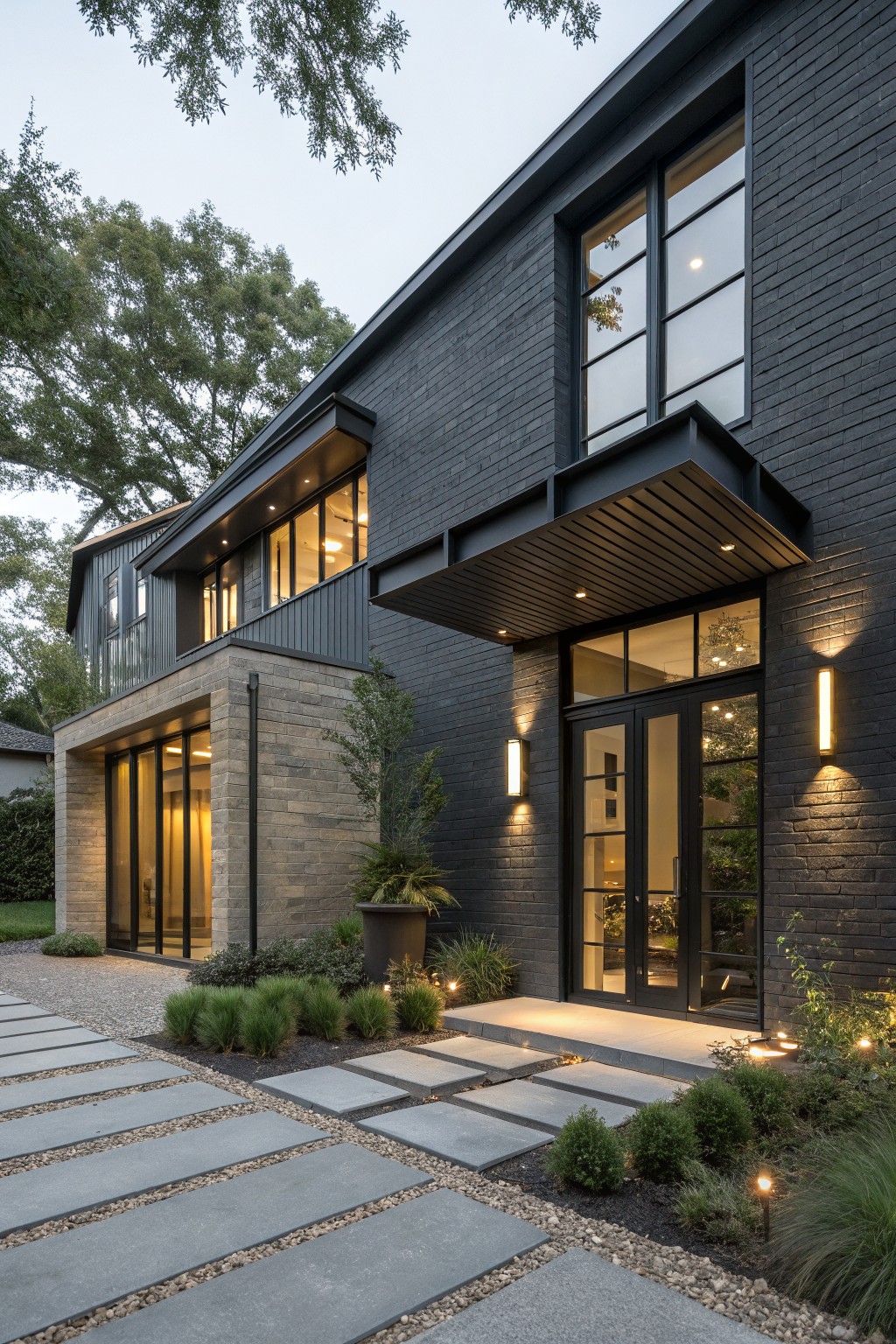 Side view of a two-story modern house with dark brick walls, a protruding wood-lined steel canopy over black-framed glass double doors, stone base section, pathway, low plantings, and exterior lights.