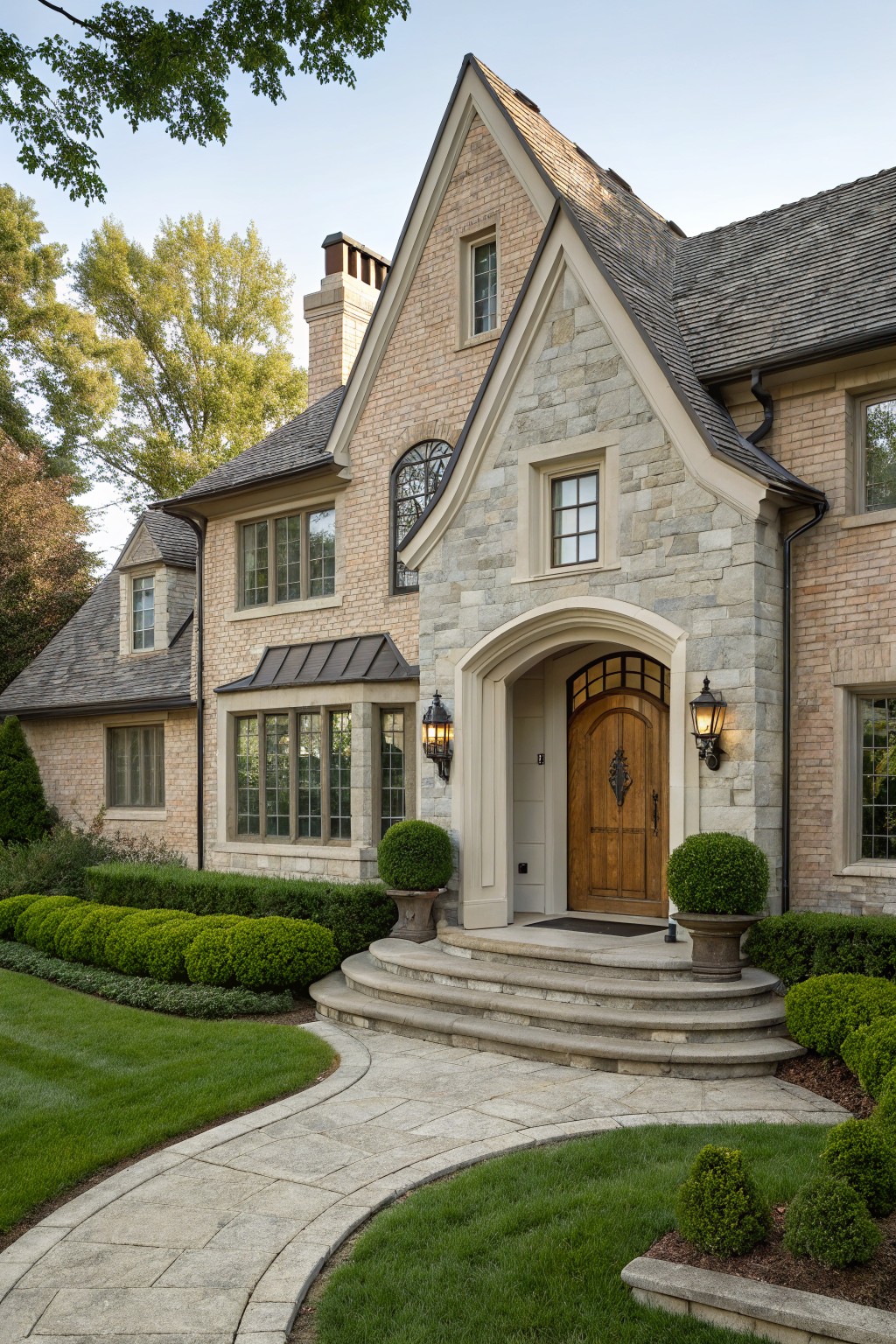 Two-story house exterior with beige brick walls, gray stone arched entryway containing a wood door with iron hardware, flanked by lanterns, wide stone steps, and low boxwood shrubs along a curved paver path in a lawn.