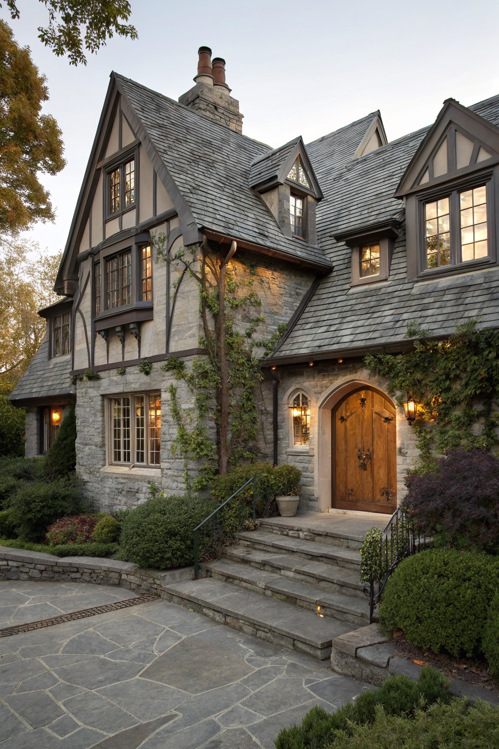 Three-story Tudor-style house exterior featuring dark half-timber framing on light stone walls, gray slate roof, arched wooden entry door with lanterns, stone steps, and ivy and shrubs in the landscaping.
