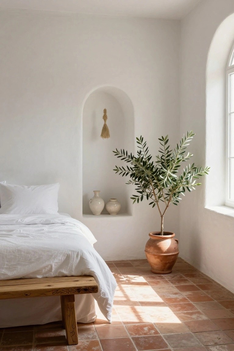 A minimalist bedroom with white walls, a low wooden bench bed draped in white linens, two white ceramic vases in a wall niche, a potted olive tree, a hanging wheat tassel, arched niche and window, and terracotta tile floor.