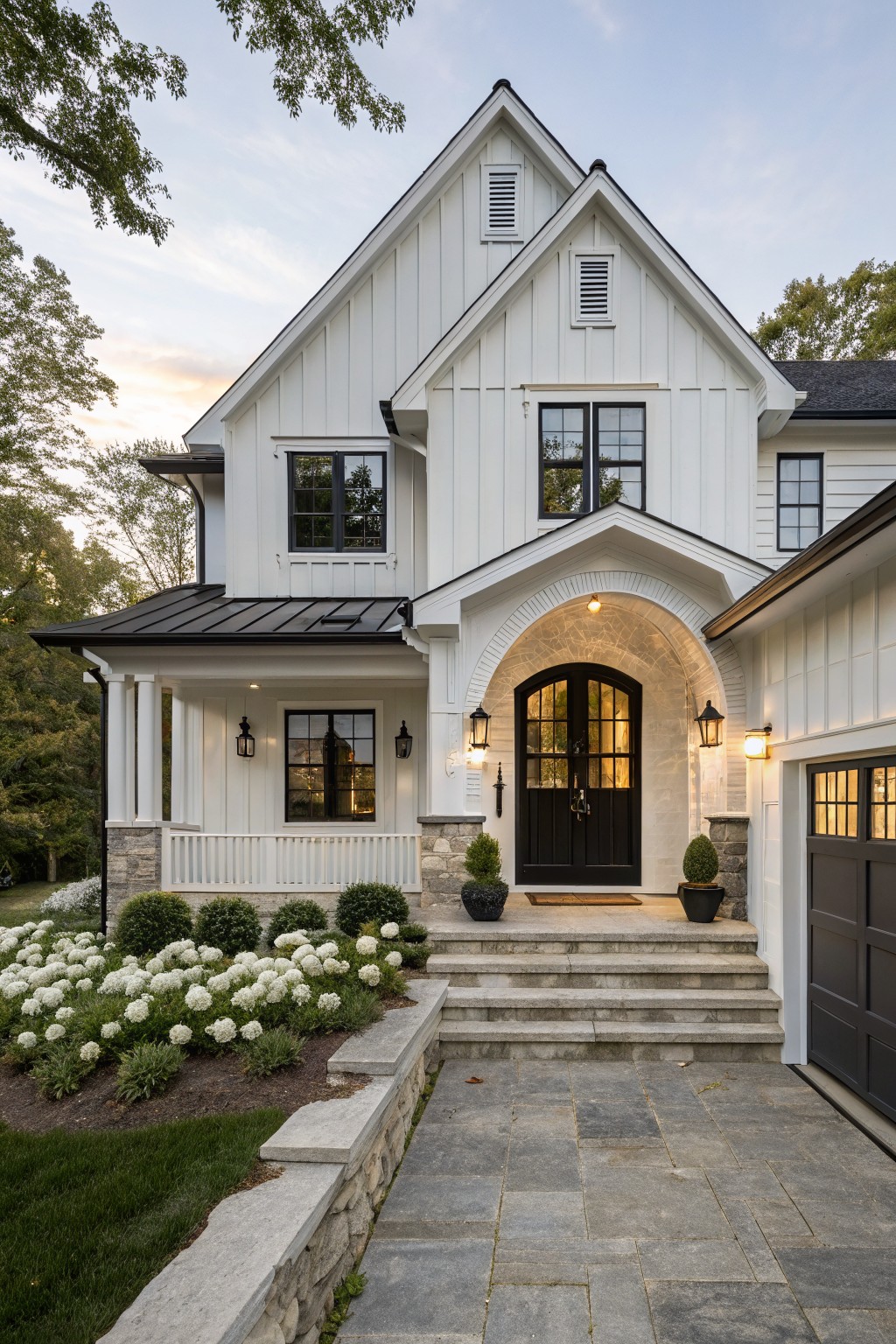White board and batten modern farmhouse exterior with black metal roof accents, arched stone entry porch featuring black double doors and lanterns, stone steps, boxwood shrubs, white hydrangea plantings, and attached garage.