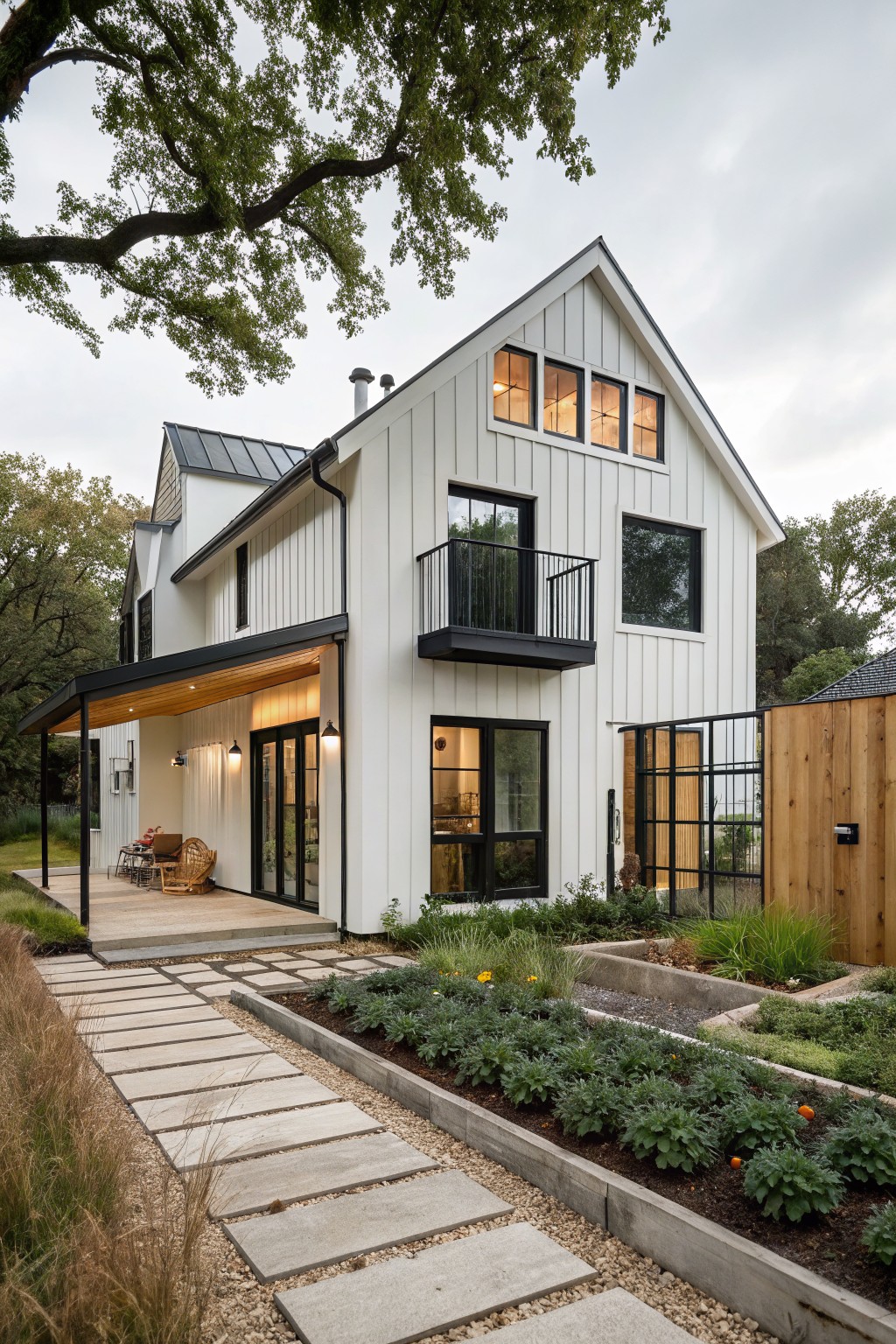 Two-story white board and batten house exterior with black metal balcony railing, covered porch, large black-framed windows, stone pathway, and raised garden beds with plants.