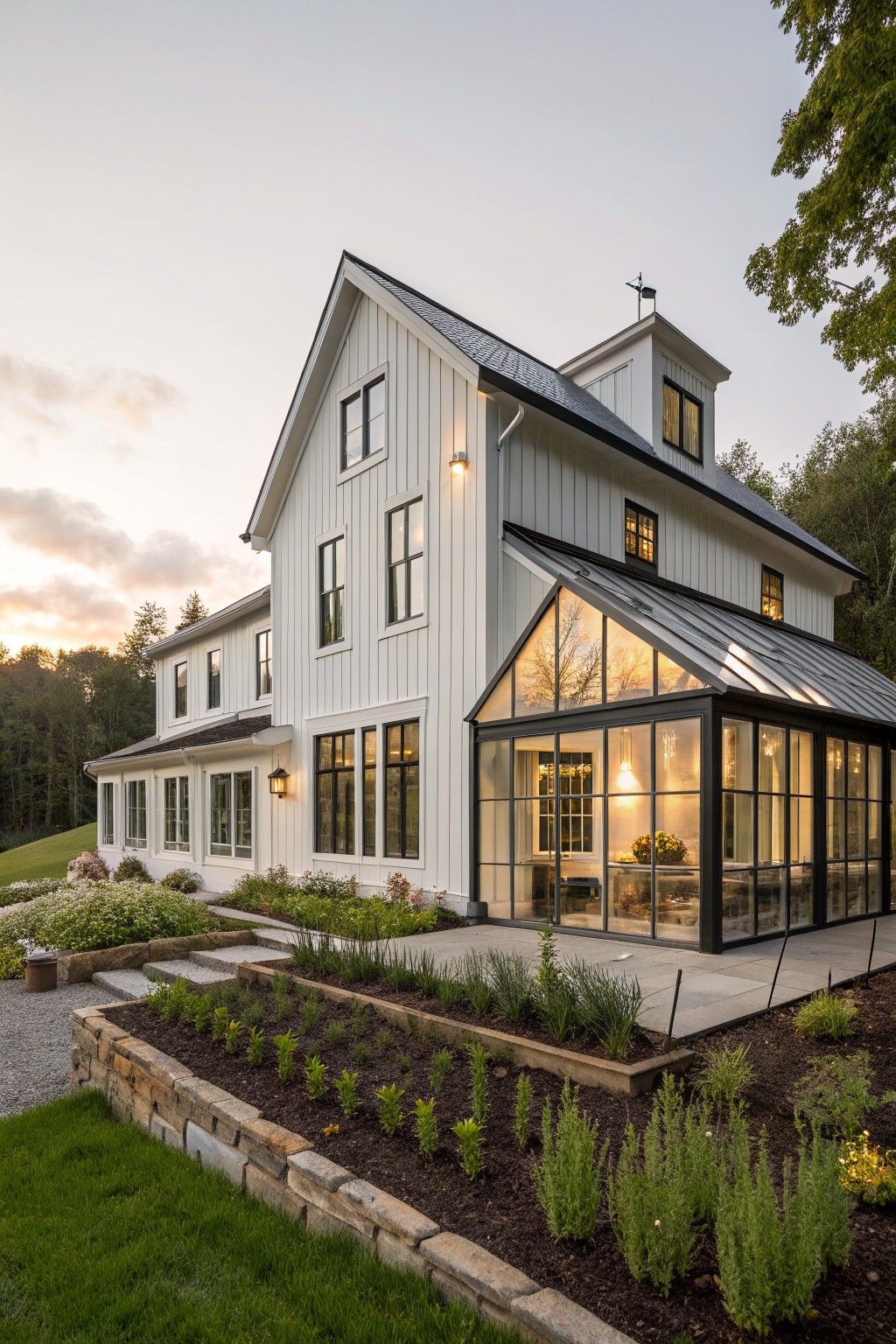 White board and batten farmhouse exterior with black metal-framed glass sunroom extension, stone steps, raised herb garden beds, gravel path, and surrounding trees at dusk.