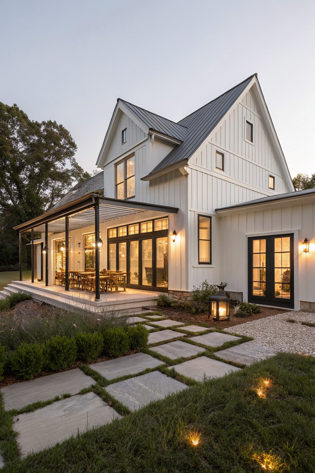 White board and batten sided two-story modern farmhouse with dark metal gable roof, covered porch featuring glass doors, dining table, black lanterns, stone paver path with grass inlays, shrubs, and dusk lighting.