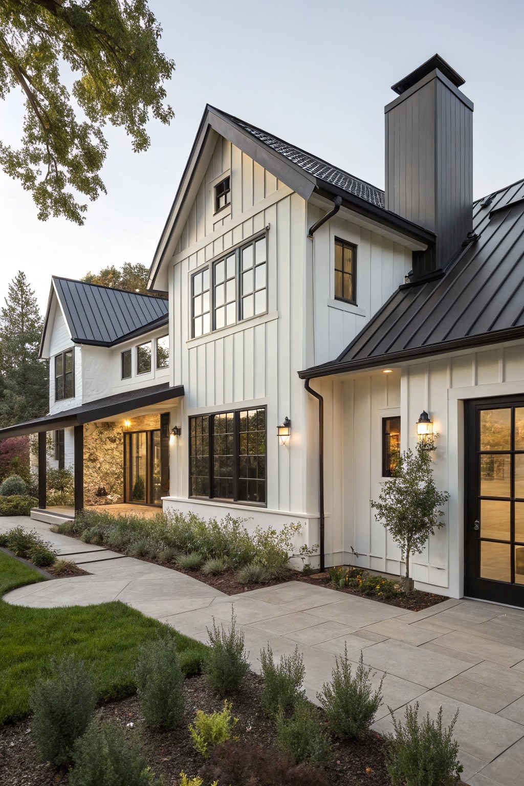 Two-story white board and batten farmhouse exterior featuring black metal roof, tall chimney, large black-framed windows, covered porch with stone accents, pathway, and low plantings.