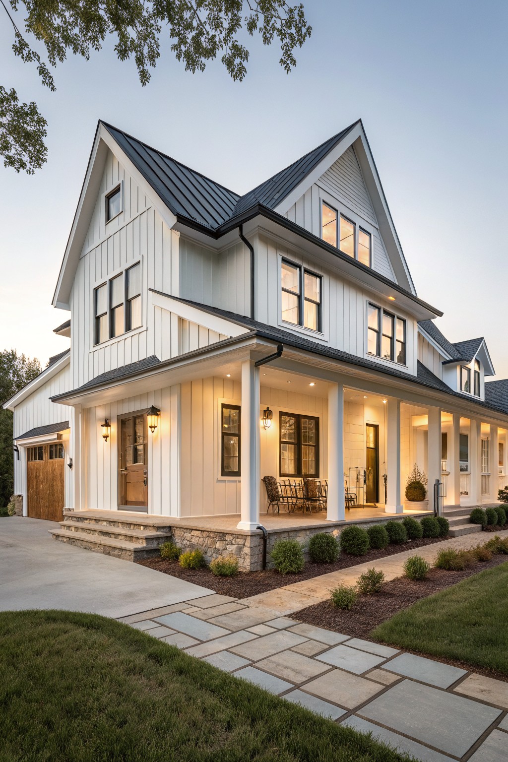 White two-story modern farmhouse exterior featuring board and batten siding, black standing seam metal roof, wraparound porch with lanterns and chairs, wood garage door, stone steps, and low landscaping along a concrete driveway at dusk.