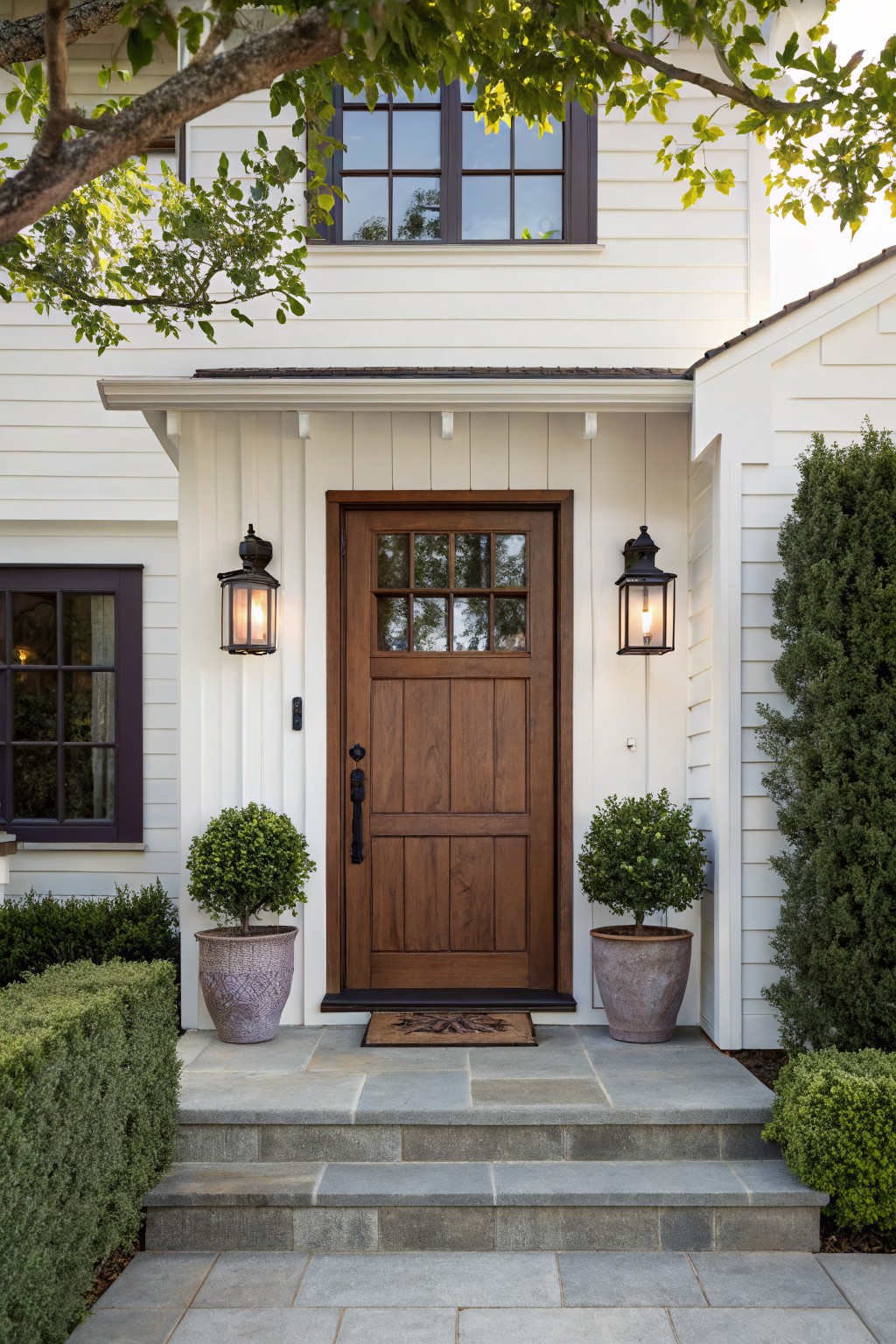White board and batten siding on a house exterior with a dark wood front door featuring glass panels, flanked by black lanterns and matching potted boxwood plants on stone steps.