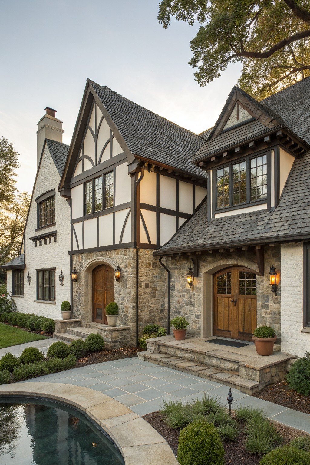 Angle view of a two-story white painted brick house with dark half-timber framing, slate roof, stone entry porch with wood door and lanterns, steps, low plants, and curved pool edge nearby.