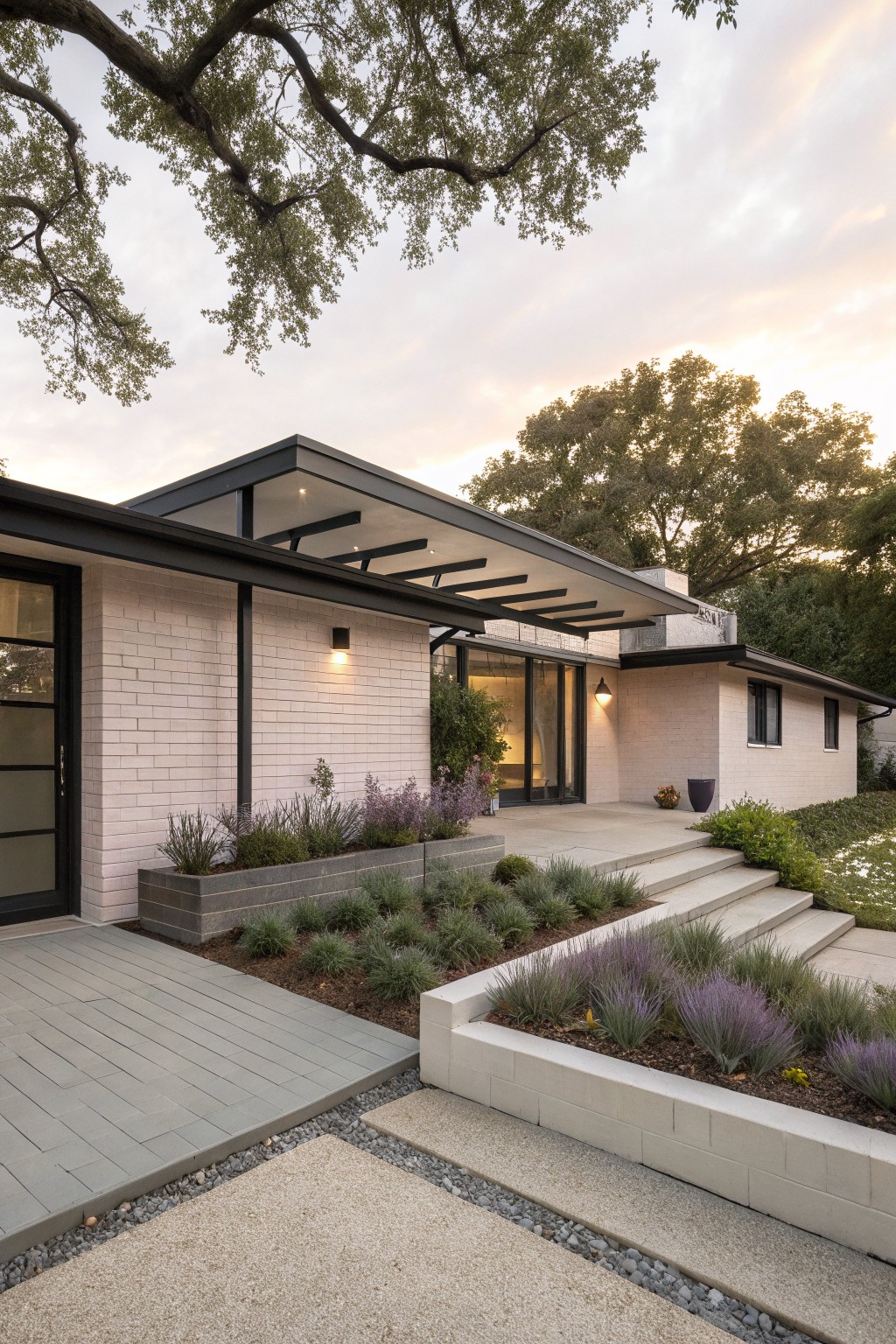 Single-story house exterior with pale painted brick walls, black metal cantilevered canopy over glass entry doors, concrete steps, raised planters with lavender, and oak trees nearby at dusk.