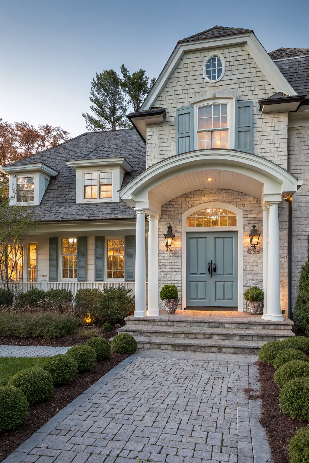 Front exterior of a two-story painted brick house with a classical portico supported by white columns, arched entryway, blue double doors, shuttered windows, and a brick pathway bordered by shrubs at dusk.