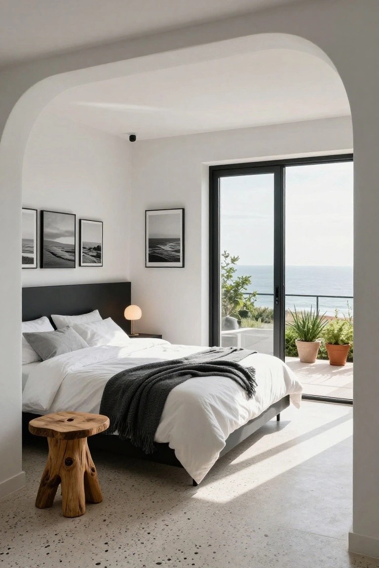 Modern bedroom with black upholstered headboard on platform bed, white bedding and pillows, black-framed black-and-white seascape photos on white walls, opal lamp on black nightstand, tree stump stool, large black sliding doors to sea-view balcony with potted plants, arched white walls, and speckled terrazzo floor.
