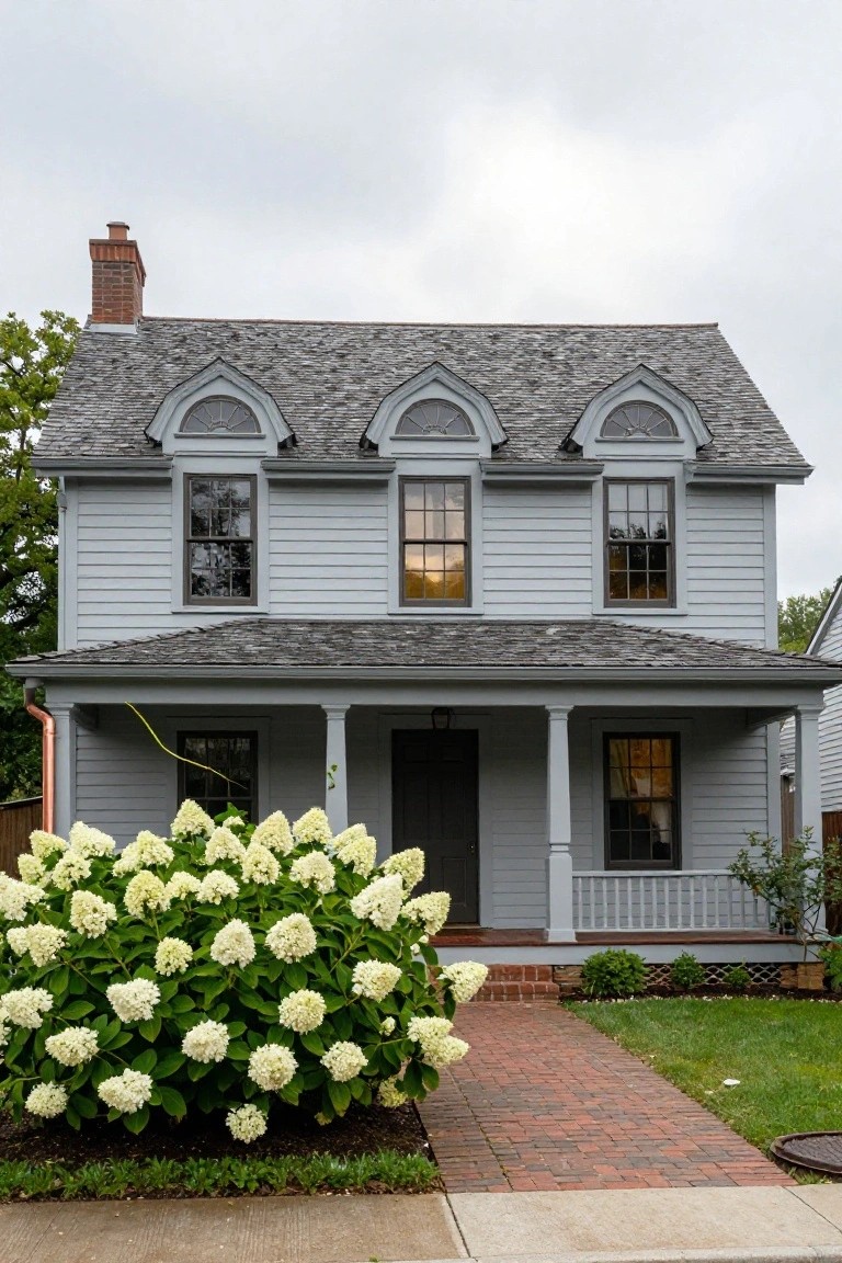Gray clapboard two-story colonial house with paired arched dormer windows on the roof, front porch with columns, dark wood door, large white hydrangea bush beside a brick walkway, and downspout along the side.