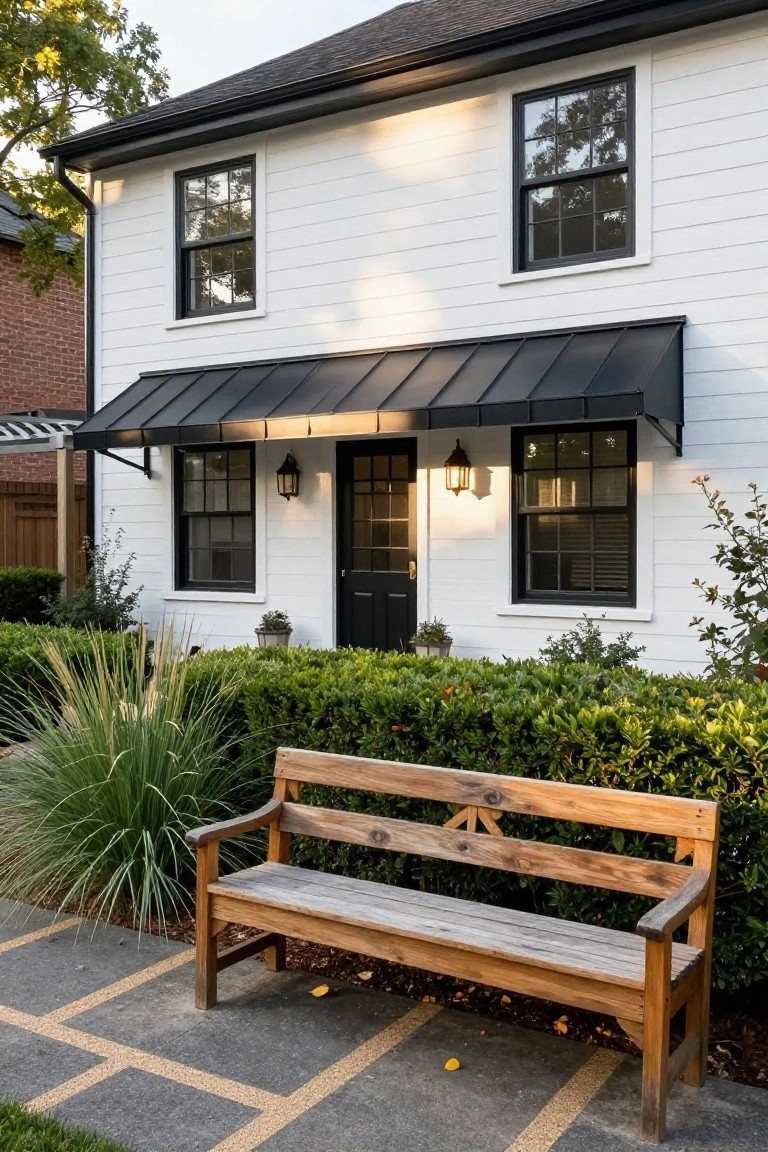 White board-and-batten house exterior with black-framed double-hung windows, black front door under a flat black metal awning supported by brackets, flanked by black lanterns, boxwood hedges, potted plants, tall grasses, and a wooden bench on a paved walkway.