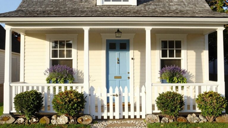 A small two-story cottage with pale yellow clapboard siding, sky blue front door with brass knocker, white trim on windows with lavender planters, white picket fence gate, boxwood shrubs, gravel path, and stone borders.