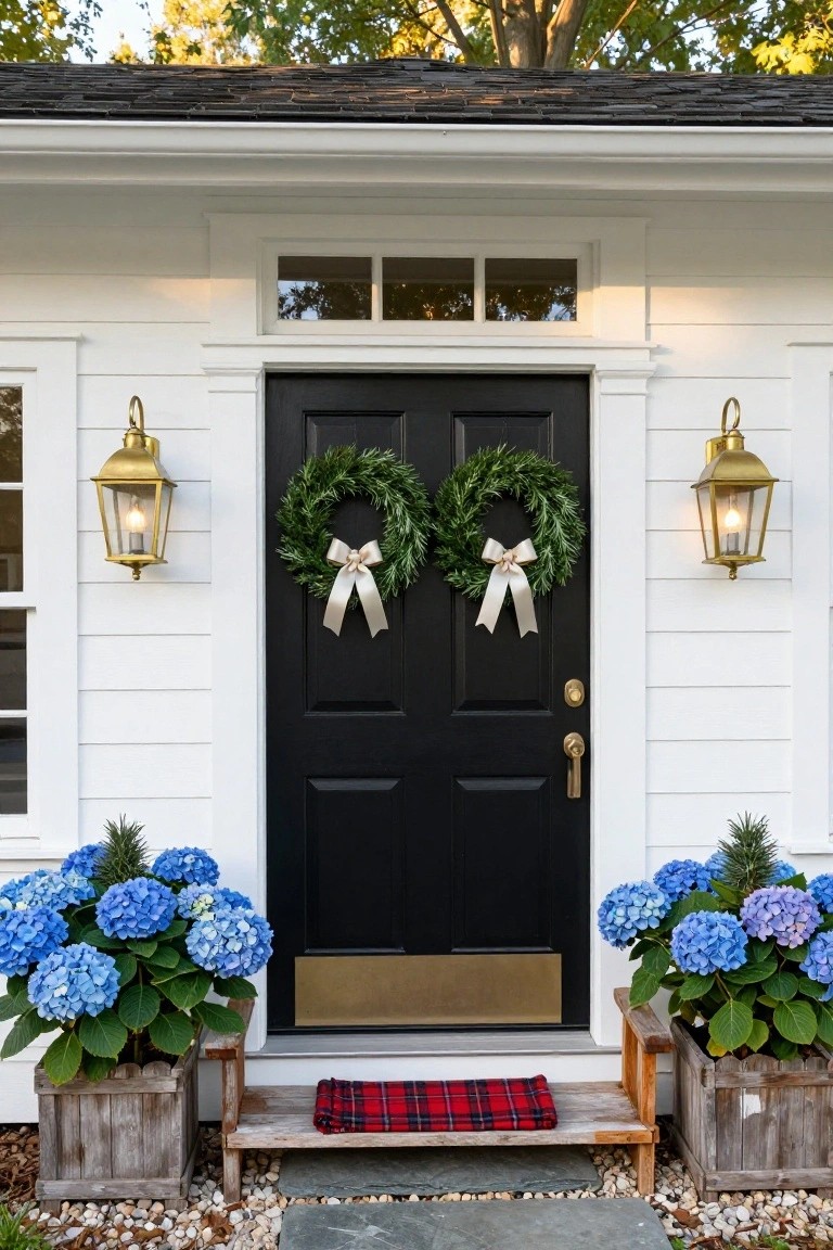 White clapboard shed with black double front doors decorated with two green evergreen wreaths tied with white satin bows, flanked by lit brass lanterns, blue hydrangea plants in wooden boxes, and a red plaid mat on a stone step.