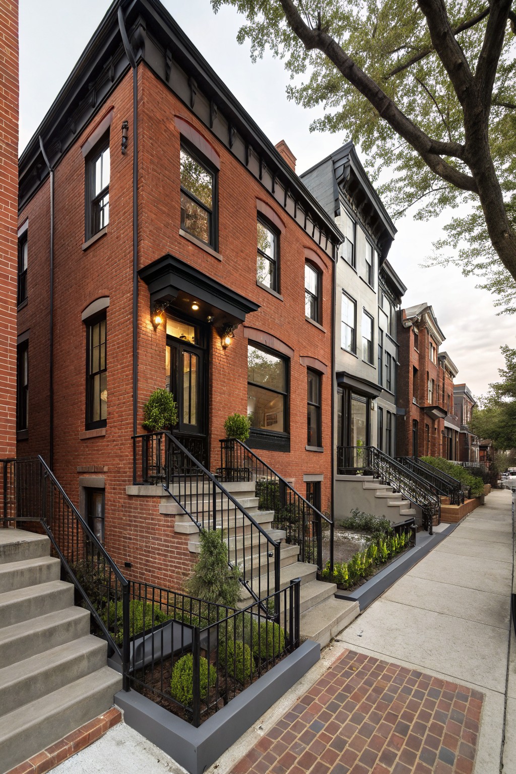 Red brick rowhouse with black trim on windows and entry porch, iron railings on concrete steps, boxwood shrubs, and adjacent townhouses along a sidewalk.