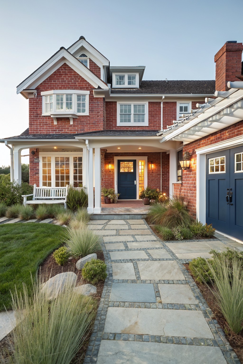 Red brick house with white trim and gabled roof, featuring a covered front porch with white columns, wooden bench, navy blue front door with glass panels, lanterns, attached garage, stone pathway, and ornamental grasses in the yard.