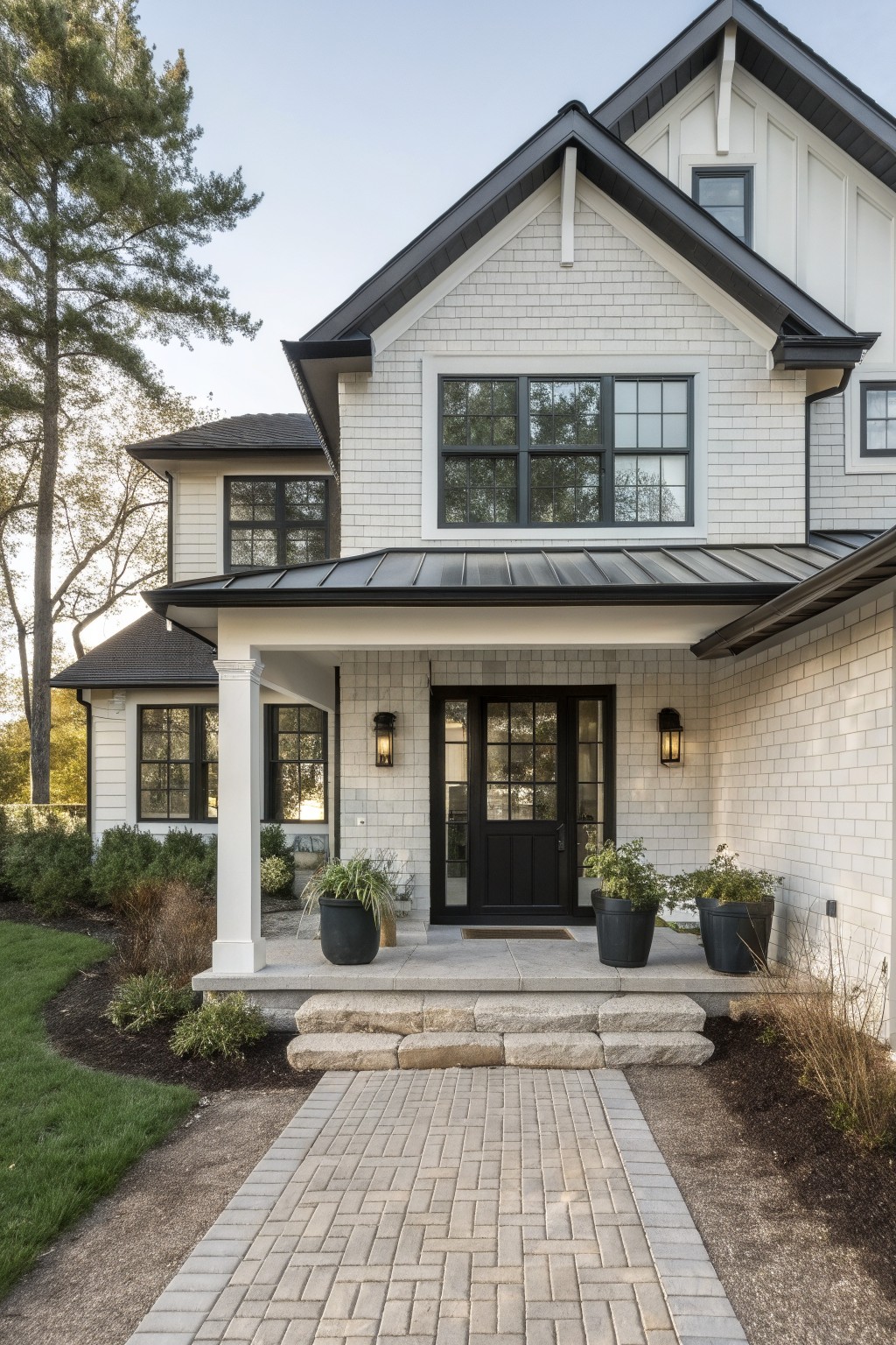 Two-story house exterior featuring light gray painted brick siding, black metal roof sections, black-trimmed windows and doors, covered front porch with stone steps and potted plants, brick pathway, and surrounding landscaping with trees and shrubs.