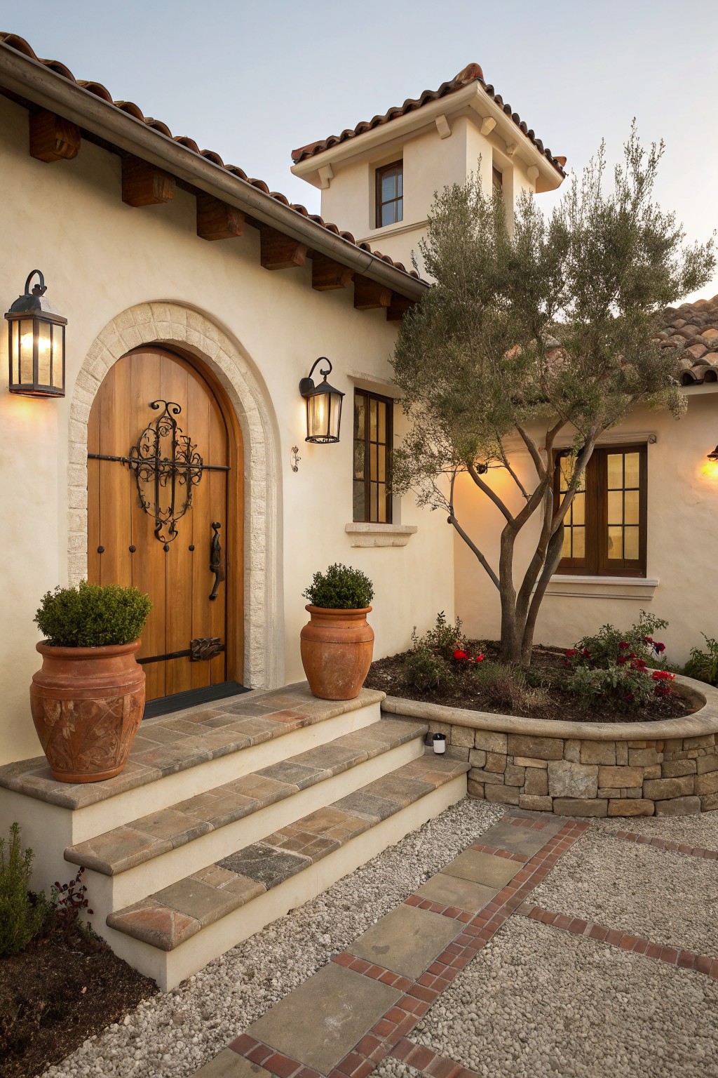 Beige stucco house exterior with arched wooden front door featuring black wrought iron crossbar and knocker, flanked by black metal lanterns and large terracotta pots on stone steps leading to a gravel path.