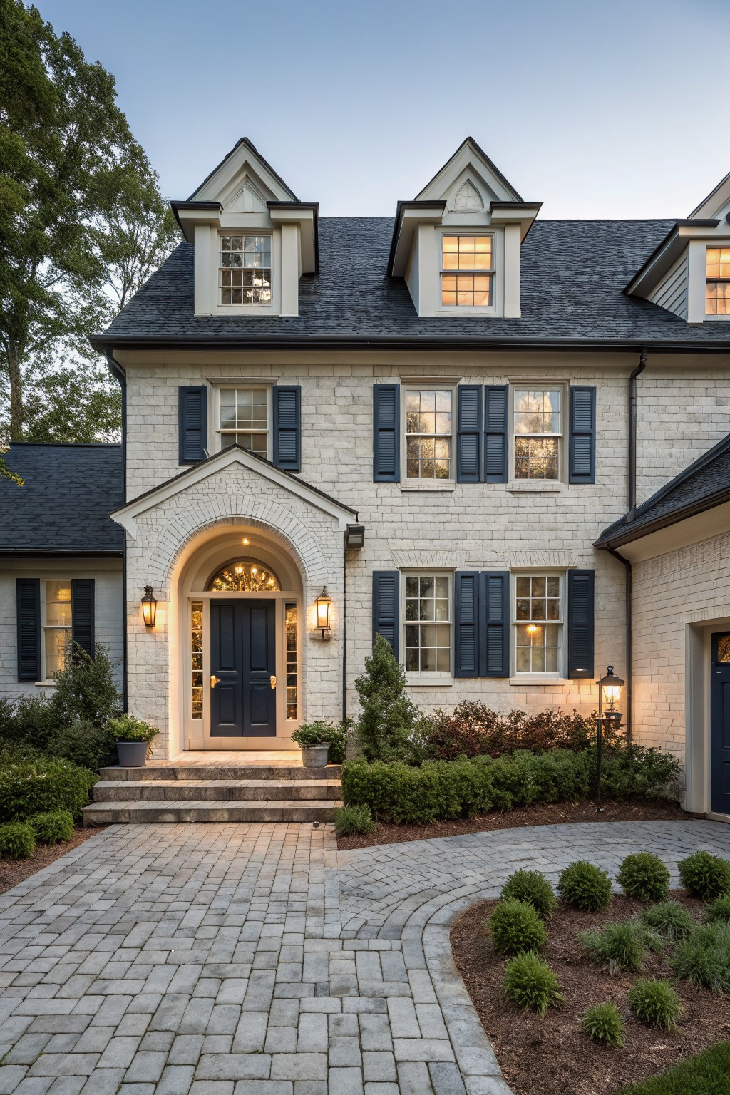 A two-story house with light painted brick exterior, navy blue shutters and double front doors, arched entryway, dark shingled roof, brick paver driveway, and low landscaping at twilight.