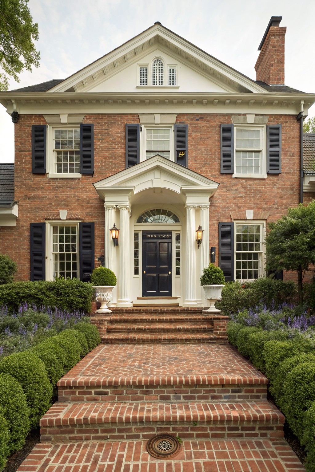 A two-story red brick house with white trim, black front door under a white portico supported by columns, flanked by lanterns, brick steps leading to the entry, boxwoods in pots, and lavender and shrubs in the front yard.