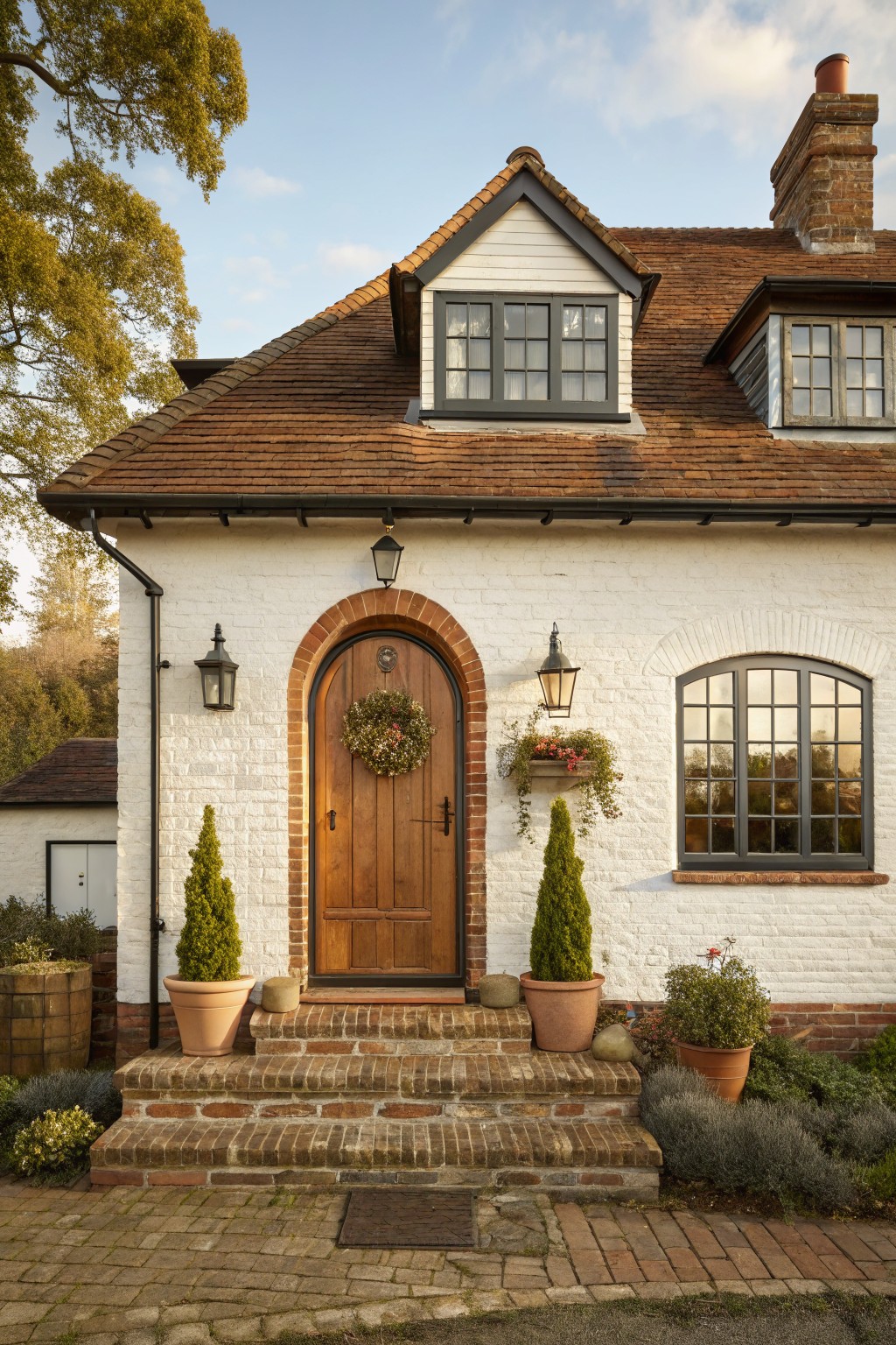 White brick house exterior with red tile roof and chimney, arched red brick entry with wooden door and wreath, flanked by lanterns and potted topiary trees, brick steps, and garden plantings along a paved path.