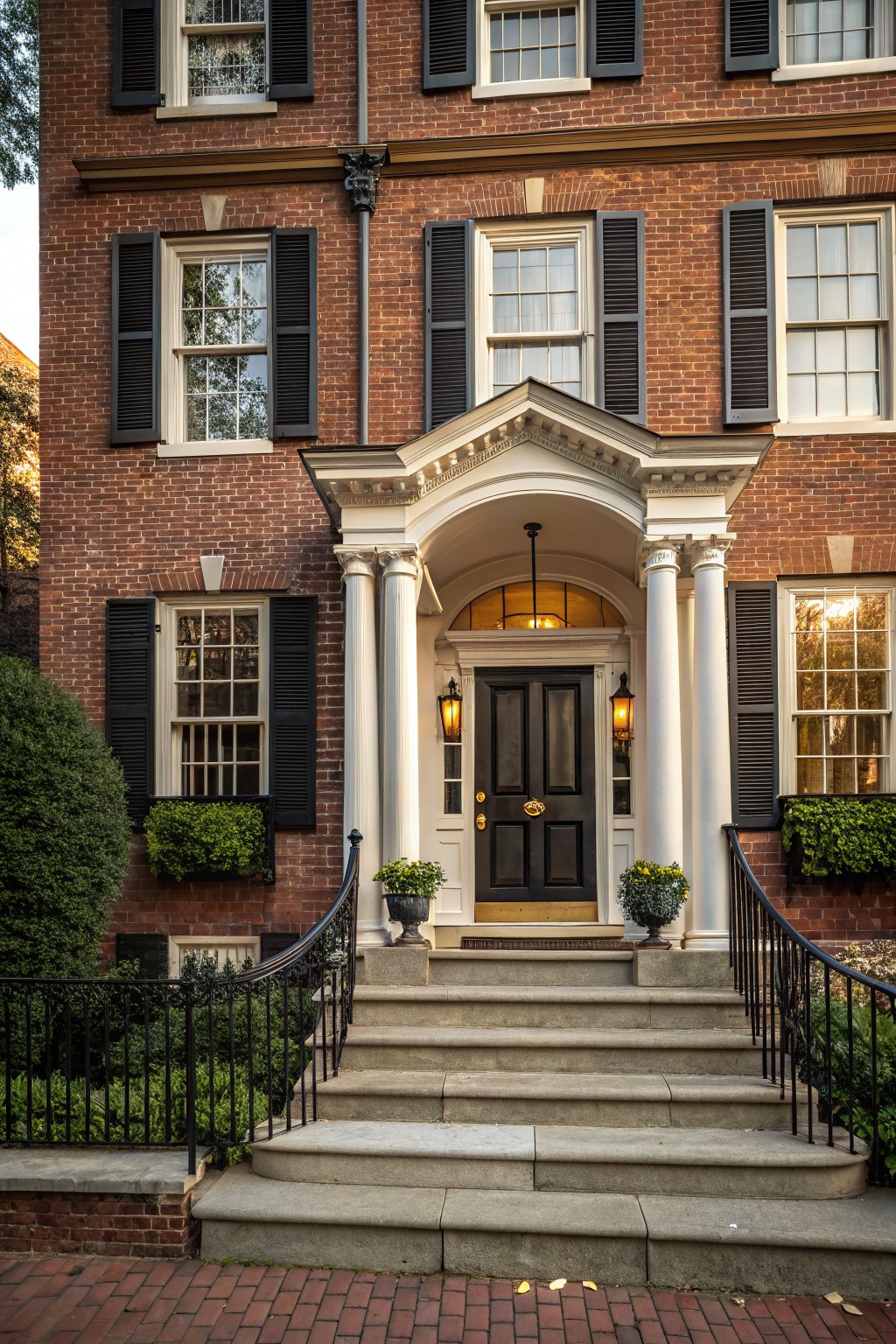Red brick townhouse with black shutters and window boxes, white columns and portico over a black front door, stone steps with black iron railings, potted plants, and brick walkway.