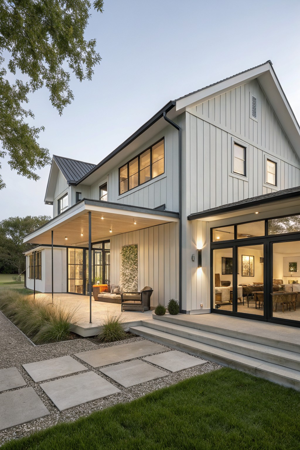 Side view of a modern farmhouse exterior with light gray board-and-batten siding, black metal roof, covered porch with lounge seating, large black-framed glass doors to interior living area, stone steps, gravel path edged by grasses, and lawn.