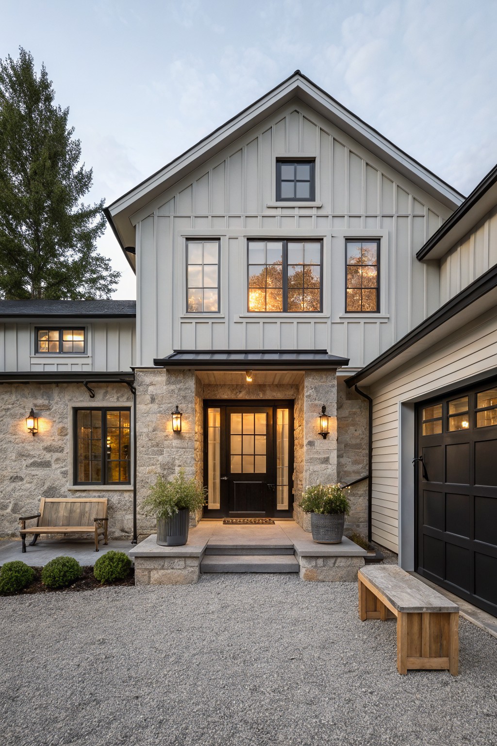 Front view of a two-story modern farmhouse with light gray board-and-batten siding, black metal roof and trim, stone entryway pillars, dark wood front door, attached garage, lanterns, wooden benches, potted plants, and gravel driveway.