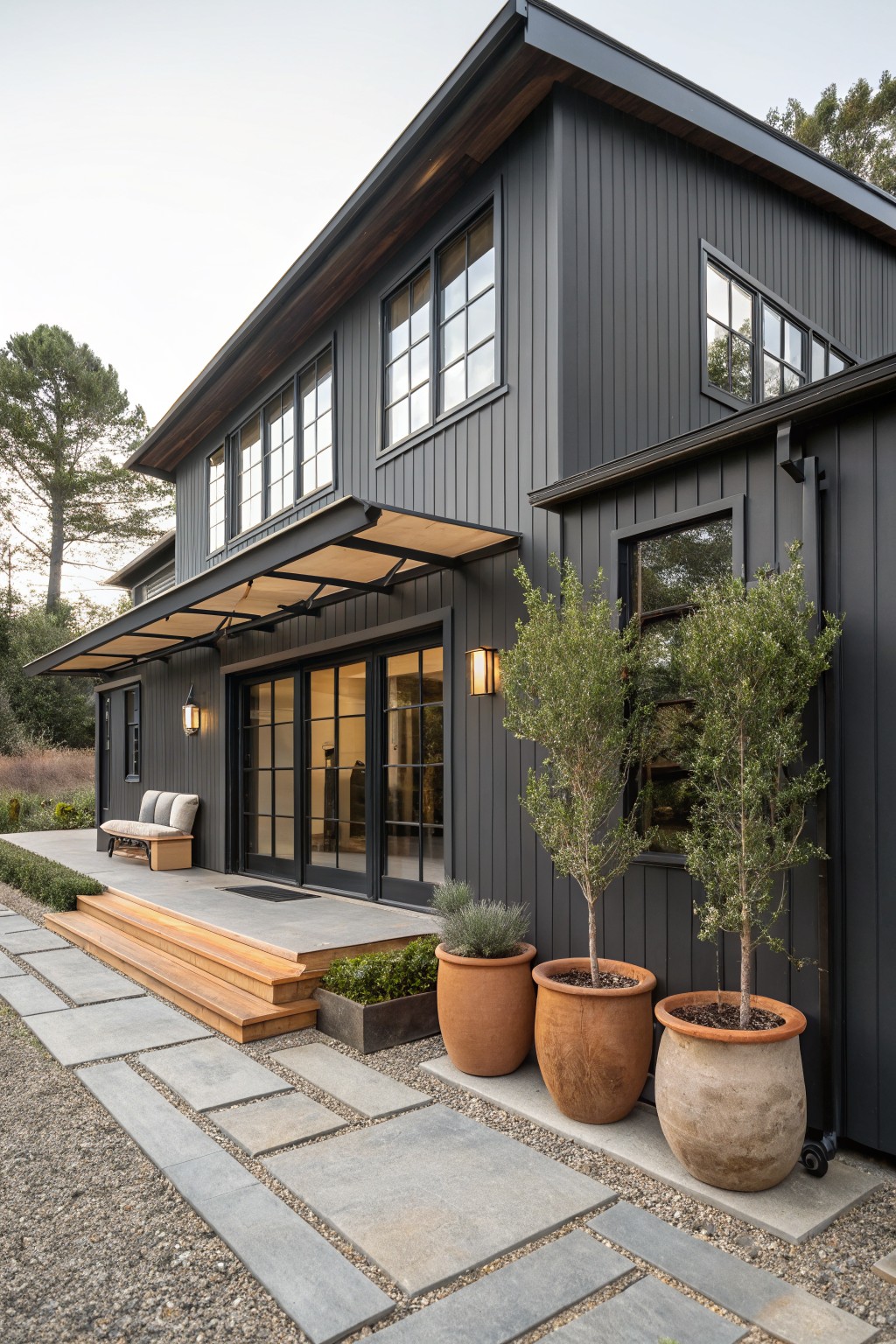 Side view of a two-story house with charcoal gray board-and-batten siding, large black-framed windows, covered entry with folding glass doors and wood steps, stone pathway, potted olive trees, and gravel ground cover.