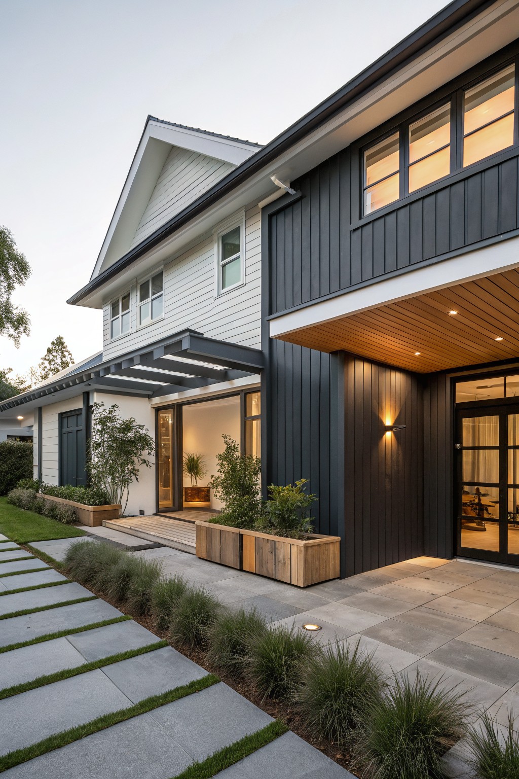 Side exterior view of a two-story modern farmhouse with white horizontal shiplap siding, dark gray vertical cladding on the entry wall, timber ceiling under the overhang, glass entry doors, concrete paver pathway, and grasses with planters.