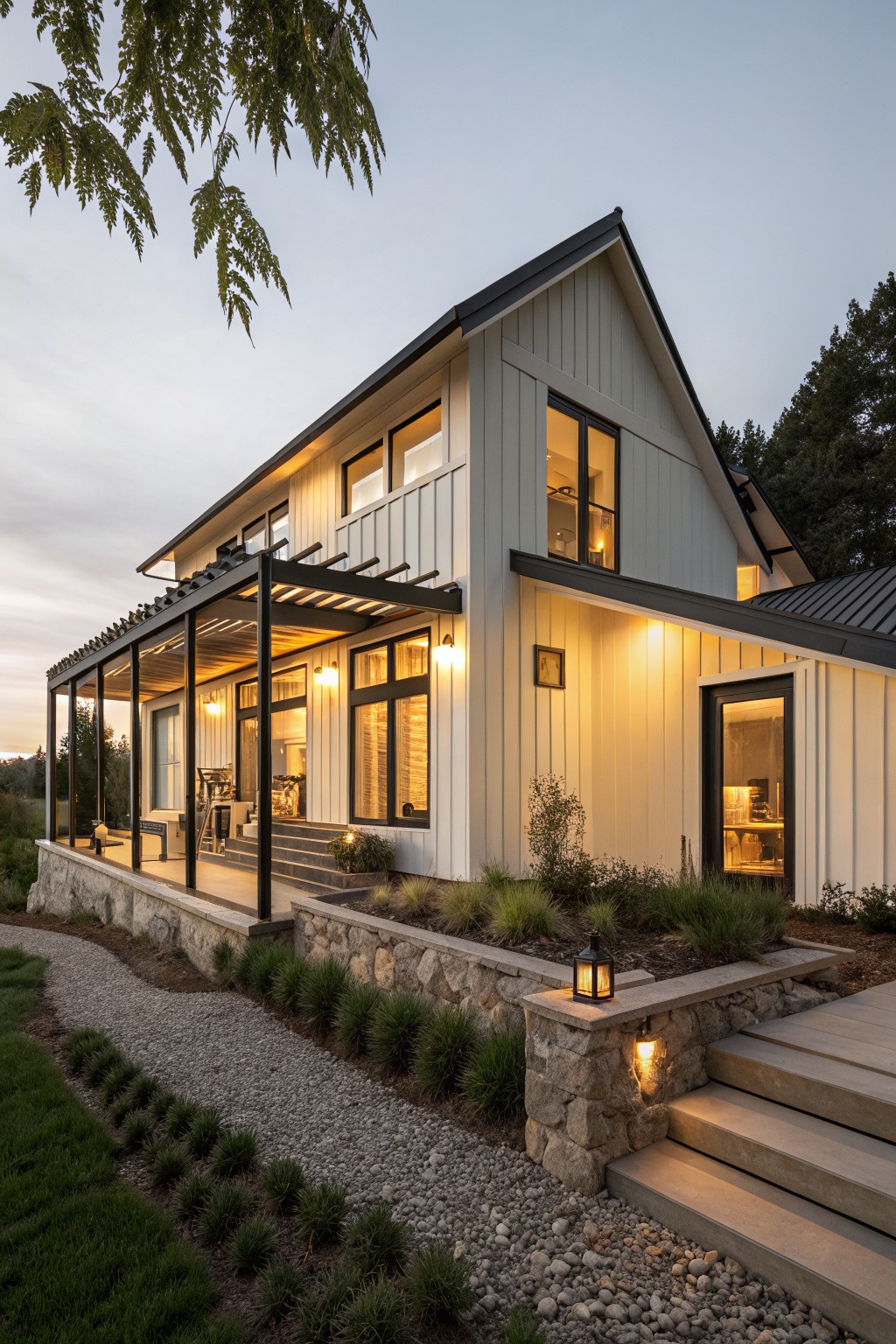 Two-story modern farmhouse exterior with light shiplap siding, black metal roof, large covered porch under black steel pergola, black-trimmed windows glowing warmly, stone retaining wall, gravel path with steps, grasses and trees at dusk.