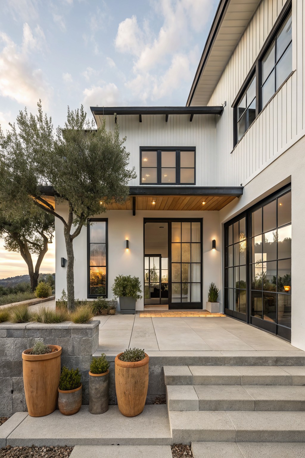 White board-and-batten house exterior with black metal-framed windows and sliding glass doors under a flat wooden overhang, flanked by an olive tree, stone steps leading to a concrete landing, and large terracotta pots with plants.