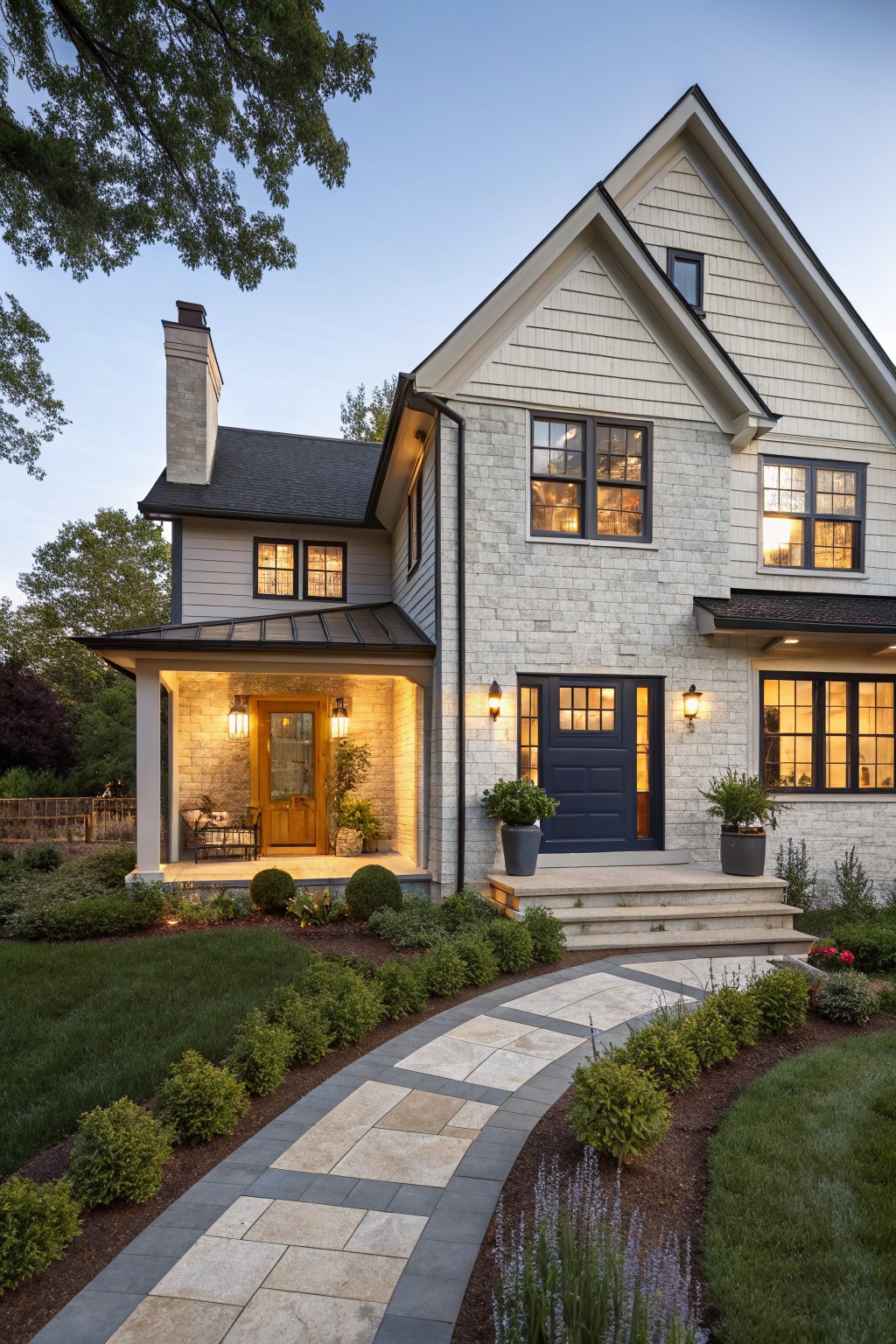 Two-story house exterior featuring light brick walls, white clapboard siding on gables, covered front porch with navy blue door, lanterns, potted plants, stone steps, and curved stone pathway edged by shrubs and grass at dusk.