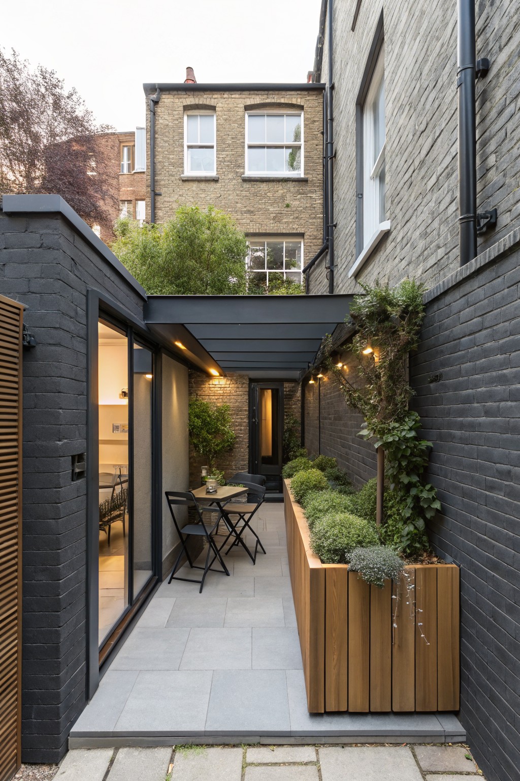 Narrow outdoor patio with gray stone pavers next to a black brick modern extension with open sliding glass doors, black metal bistro chairs and table, and a tall wooden planter box filled with greenery along the wall.