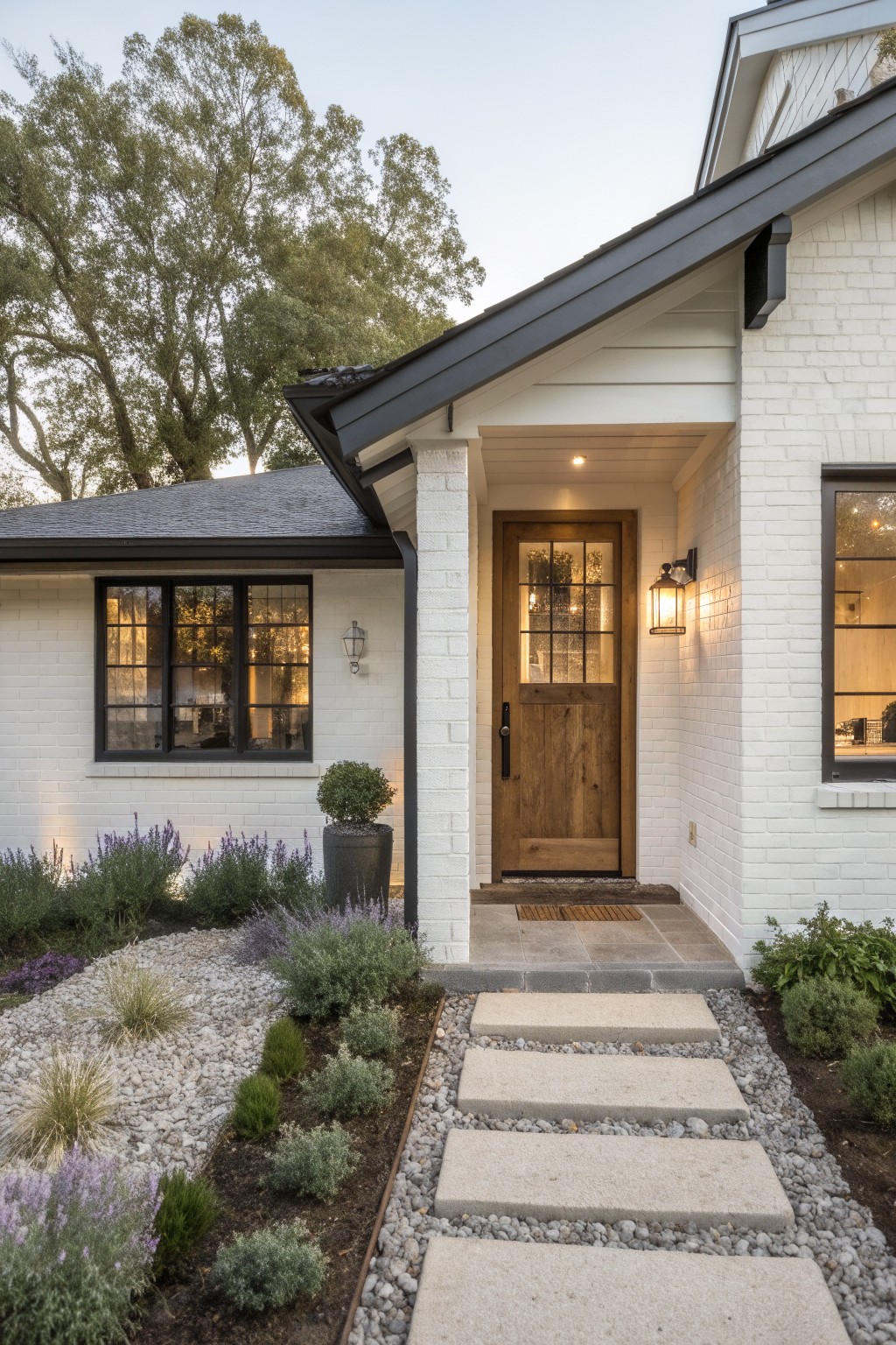 White painted brick house exterior with a dark wooden front door under a covered porch, black-framed windows, lantern lights, and a stone step path leading through gravel landscaping.