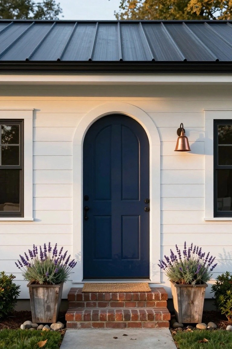 White clapboard house exterior featuring a centered navy blue arched front door with black hardware, flanked by lavender plants in rustic wooden planters, brick entry steps, copper wall lantern, black-framed windows, and dark metal roof under autumn trees.