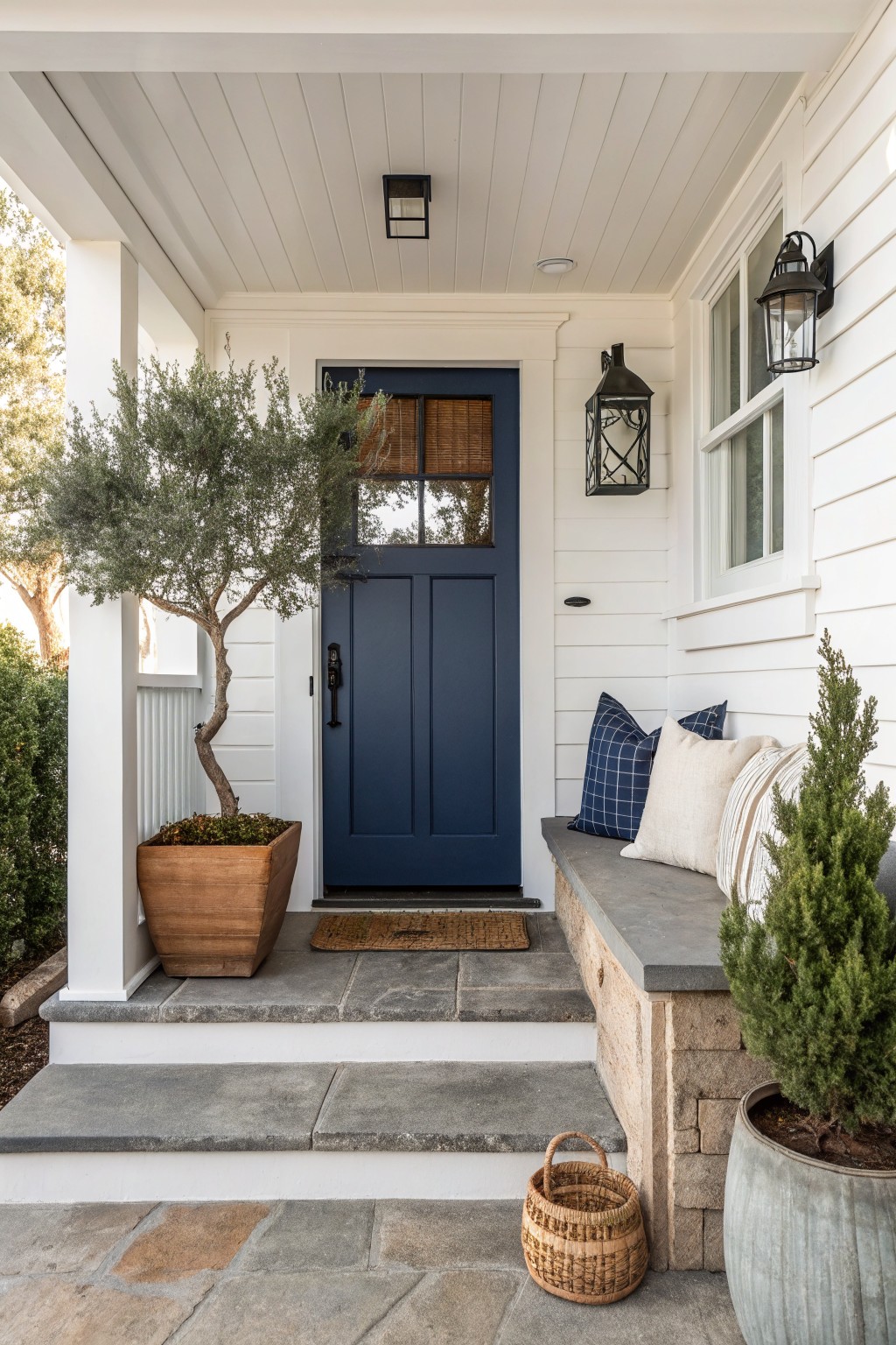 White modern farmhouse porch with navy blue double front door, flanked by lanterns and windows, olive tree in terracotta pot, stone bench with blue and white pillows, doormat, small potted evergreens, and basket on slate steps.